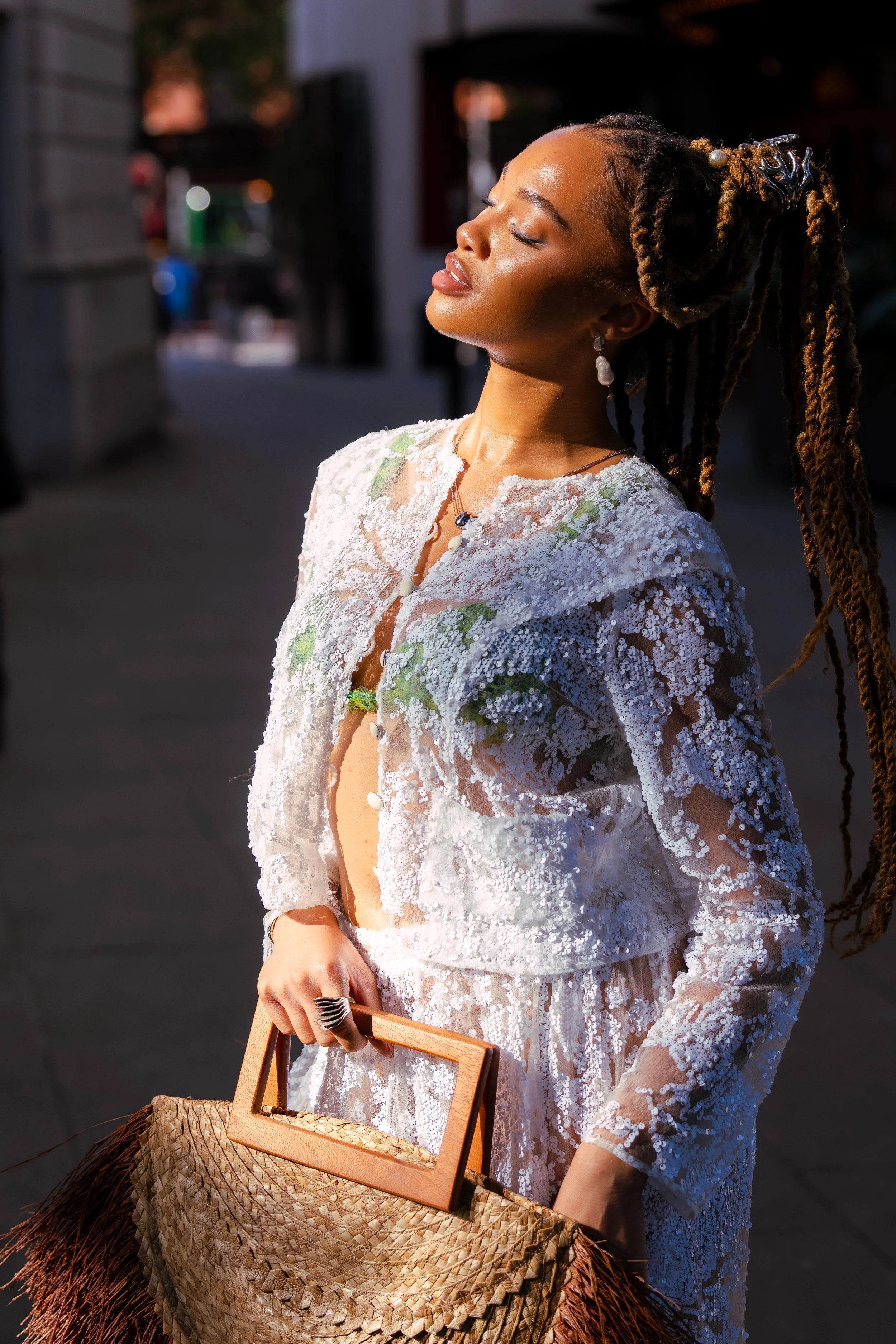 A woman with closed eyes and a serene expression, wearing a white lace outfit, standing outdoors in sunlight, holding a woven straw bag with a wooden handle.