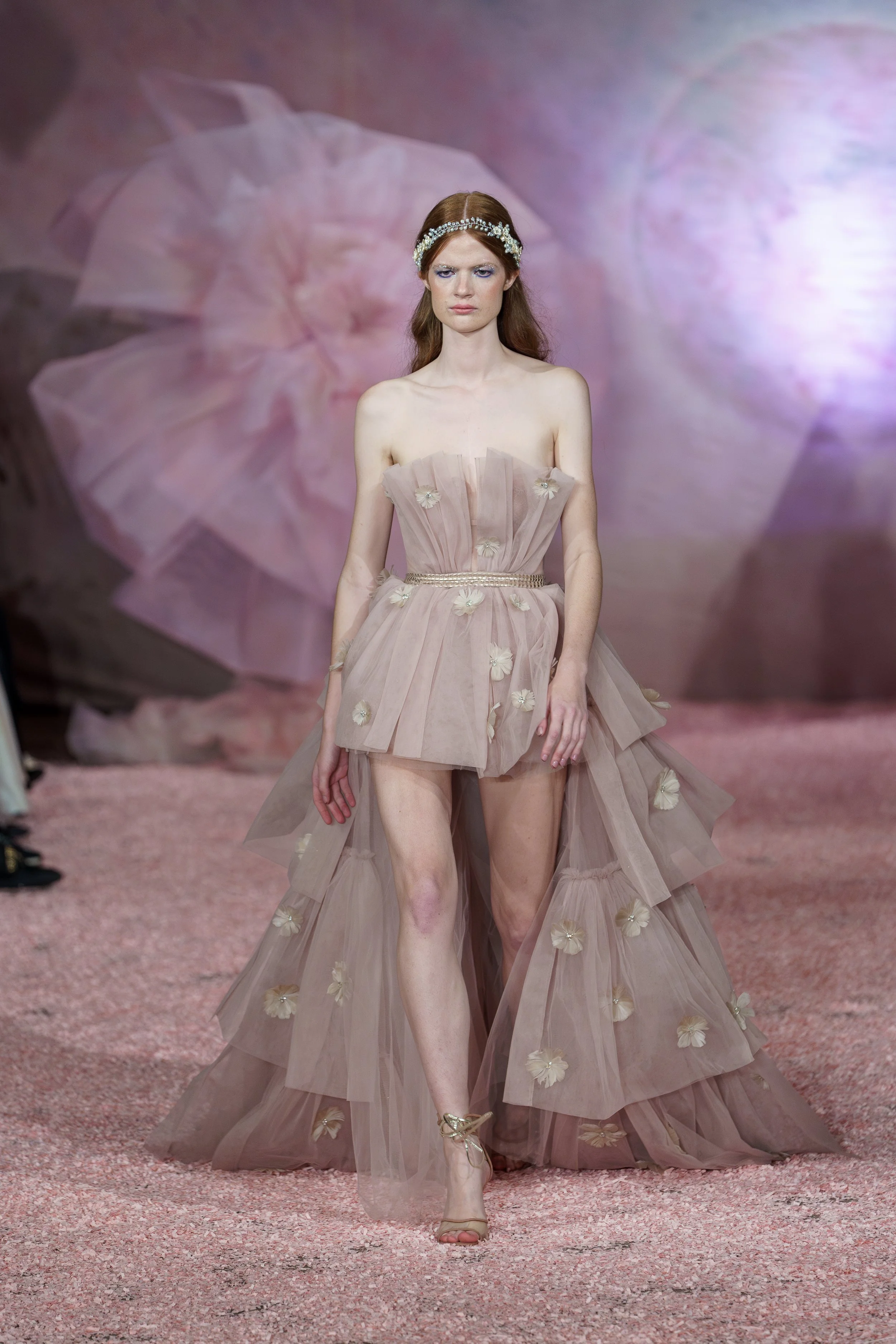 A fashion model walking down a runway wearing a sheer, layered pink dress with floral details and a matching veil, accessorized with a pearl headband and gold heels.