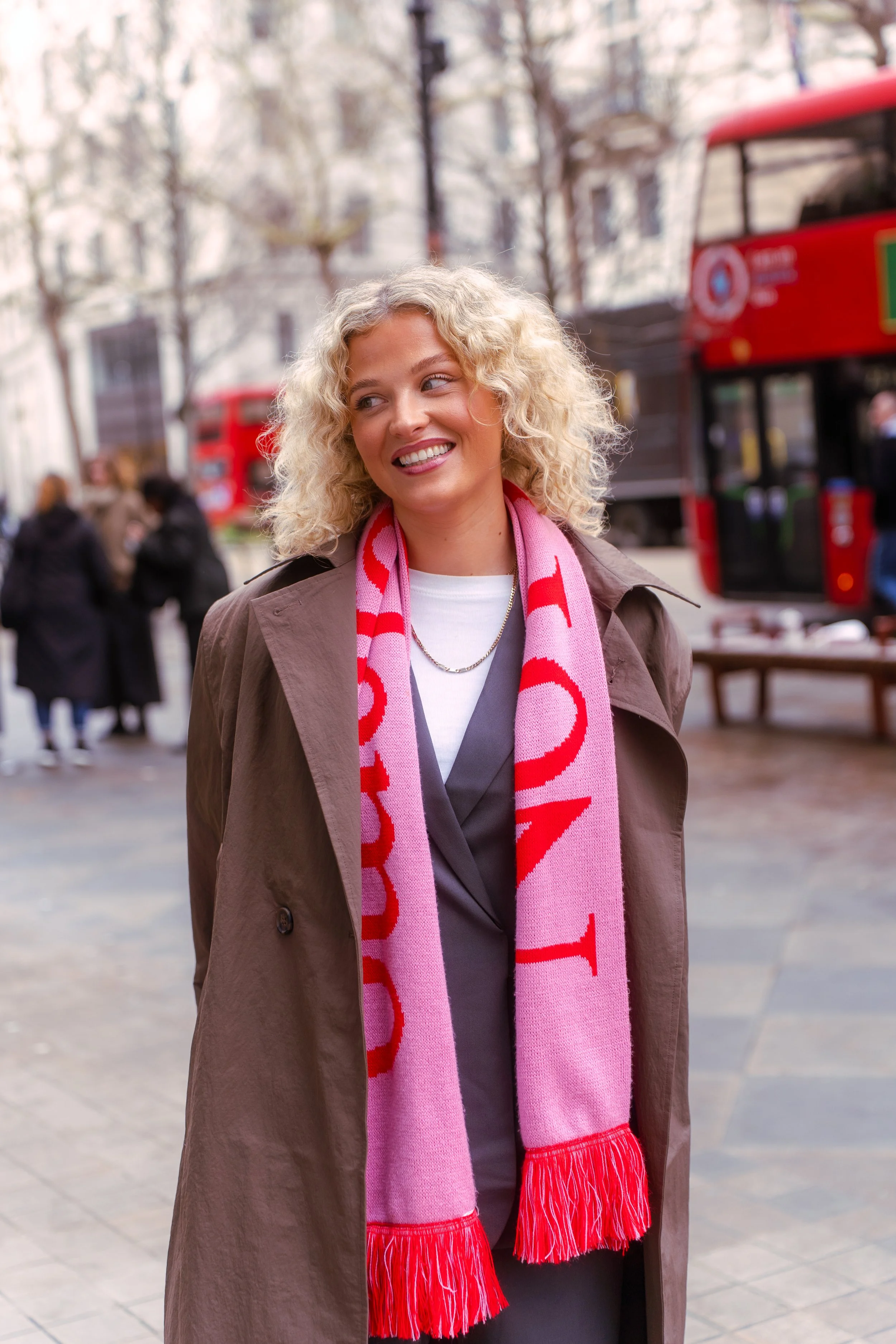 A smiling woman with curly blonde hair wearing a brown trench coat and a pink scarf, standing outdoors on a city street with red buses and people in the background.