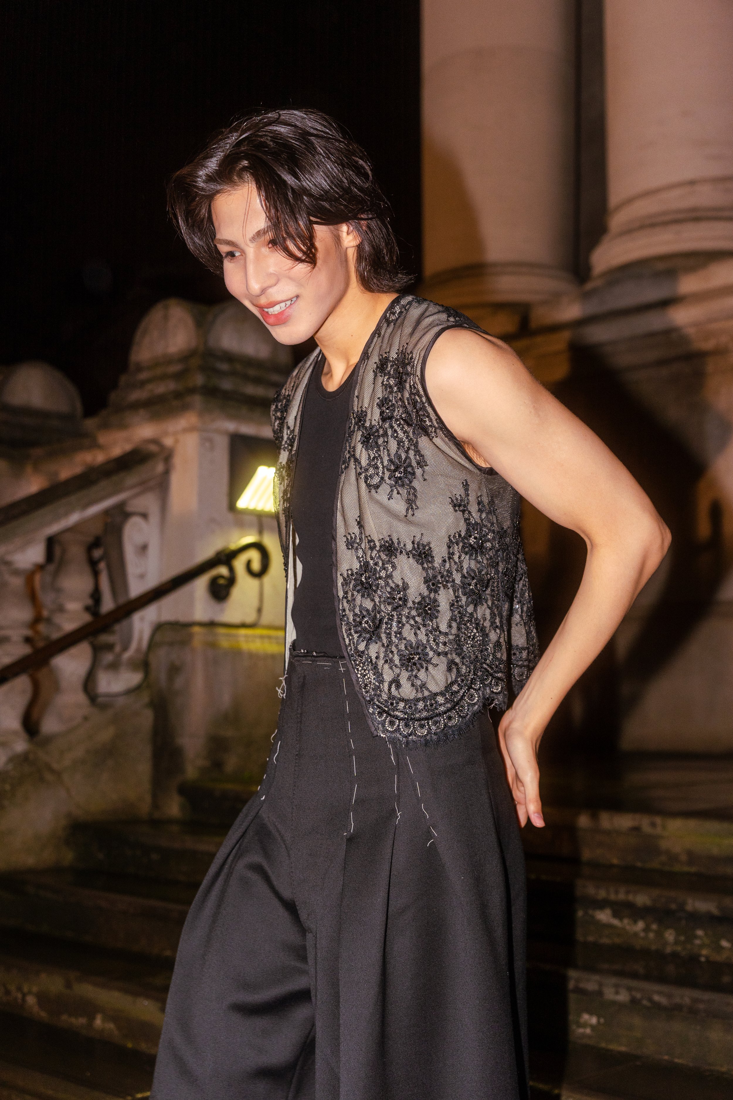 A young woman with short dark hair smiling, wearing a sleeveless black lace top and black wide-leg pants, standing outdoors at night near stone steps and columns.