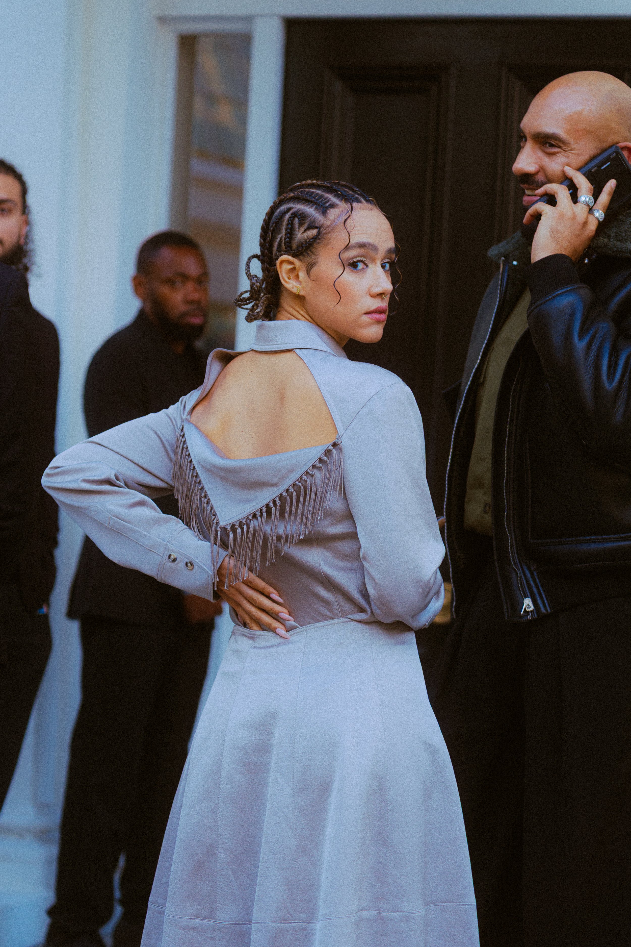 A woman with braided hair looking over her shoulder in a gray outfit, standing indoors next to a man on the phone, with two other men in the background.