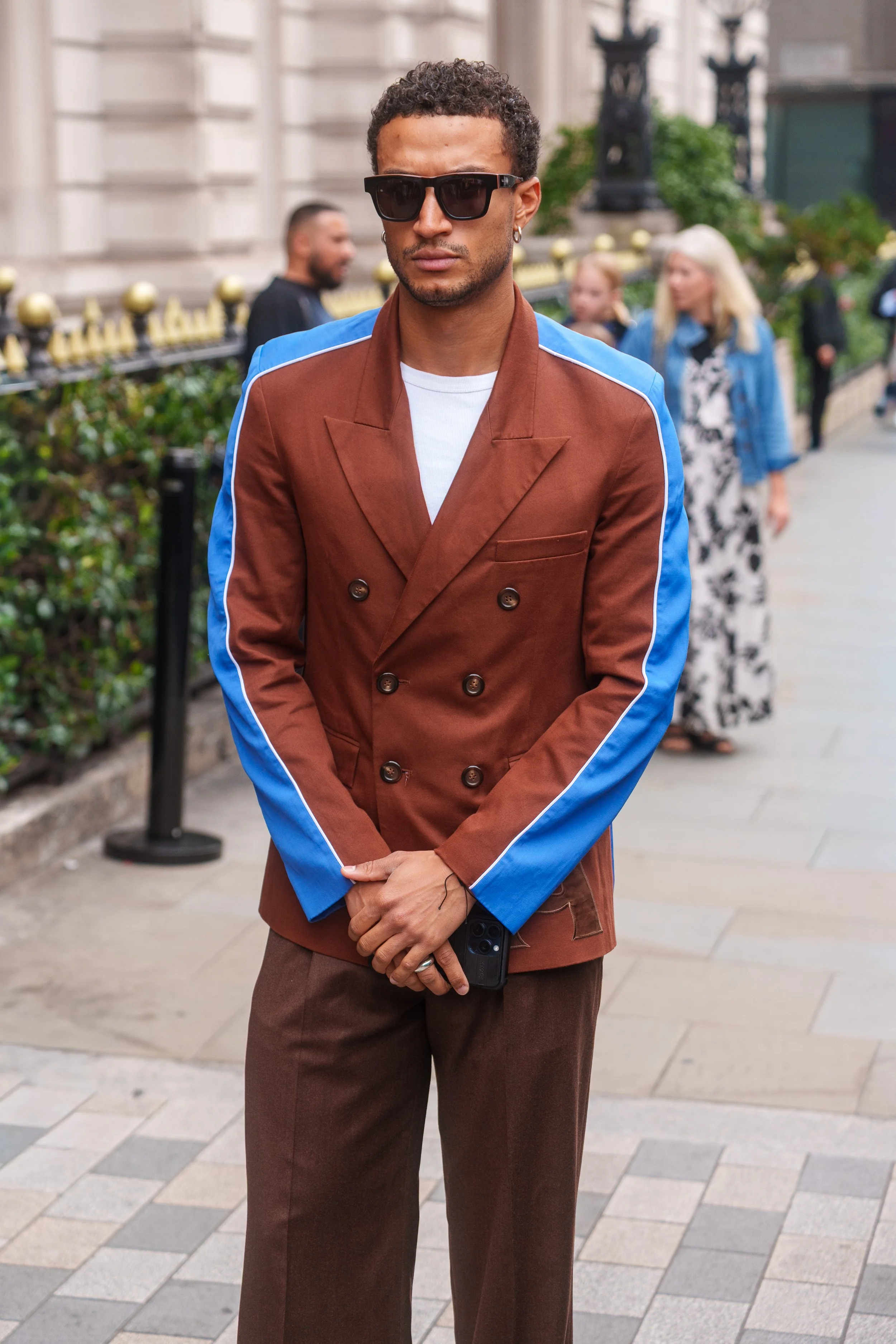 A man with curly hair and sunglasses wearing a brown double-breasted jacket with blue stripes on the sleeves and brown pants, standing on a city sidewalk.