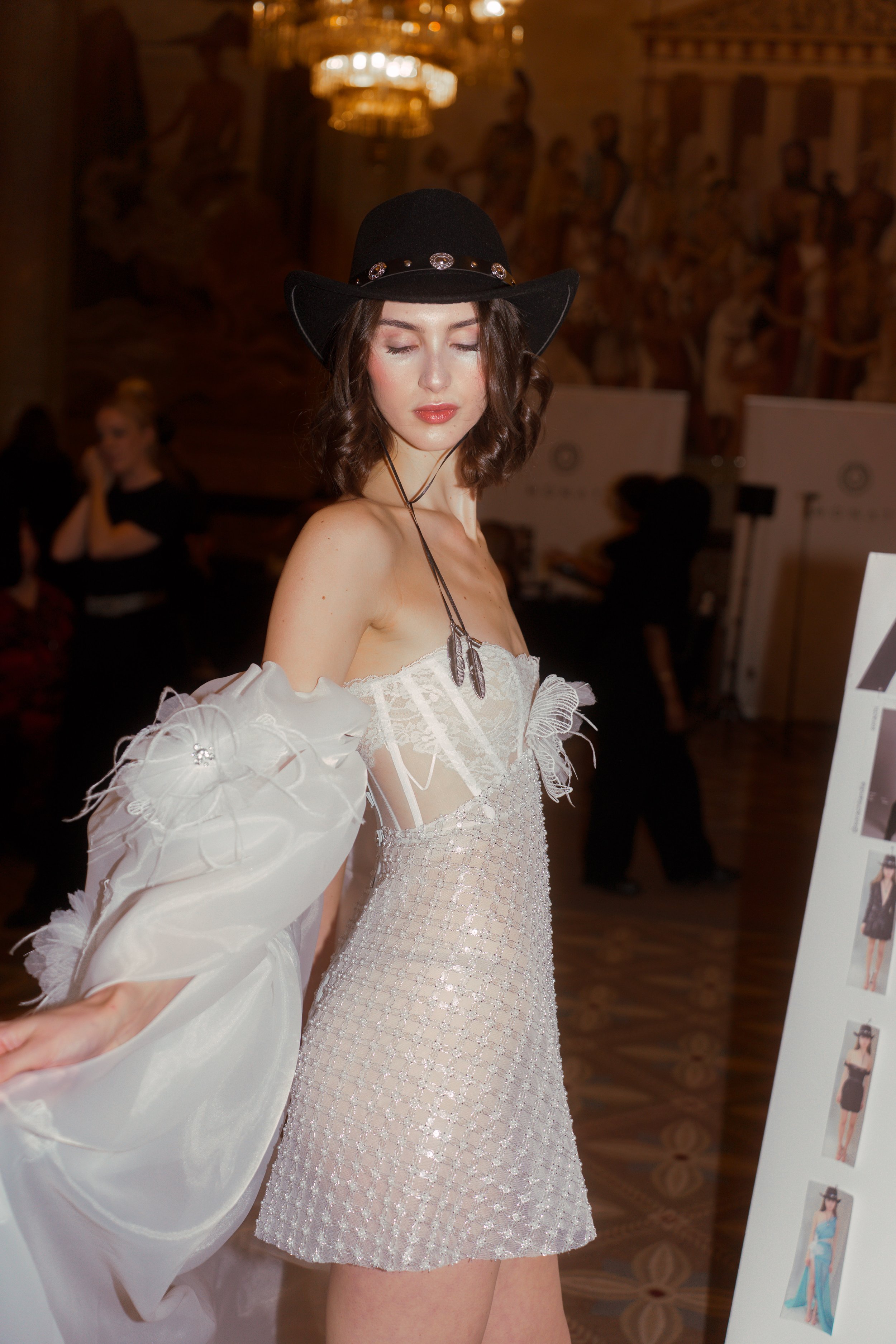 A young woman at a fashion event, wearing a black wide-brimmed hat with decorative embellishments and a strapless, white, sheer dress with ruffled sleeves, standing in a grand hall with a mural on the wall and ornate chandeliers.