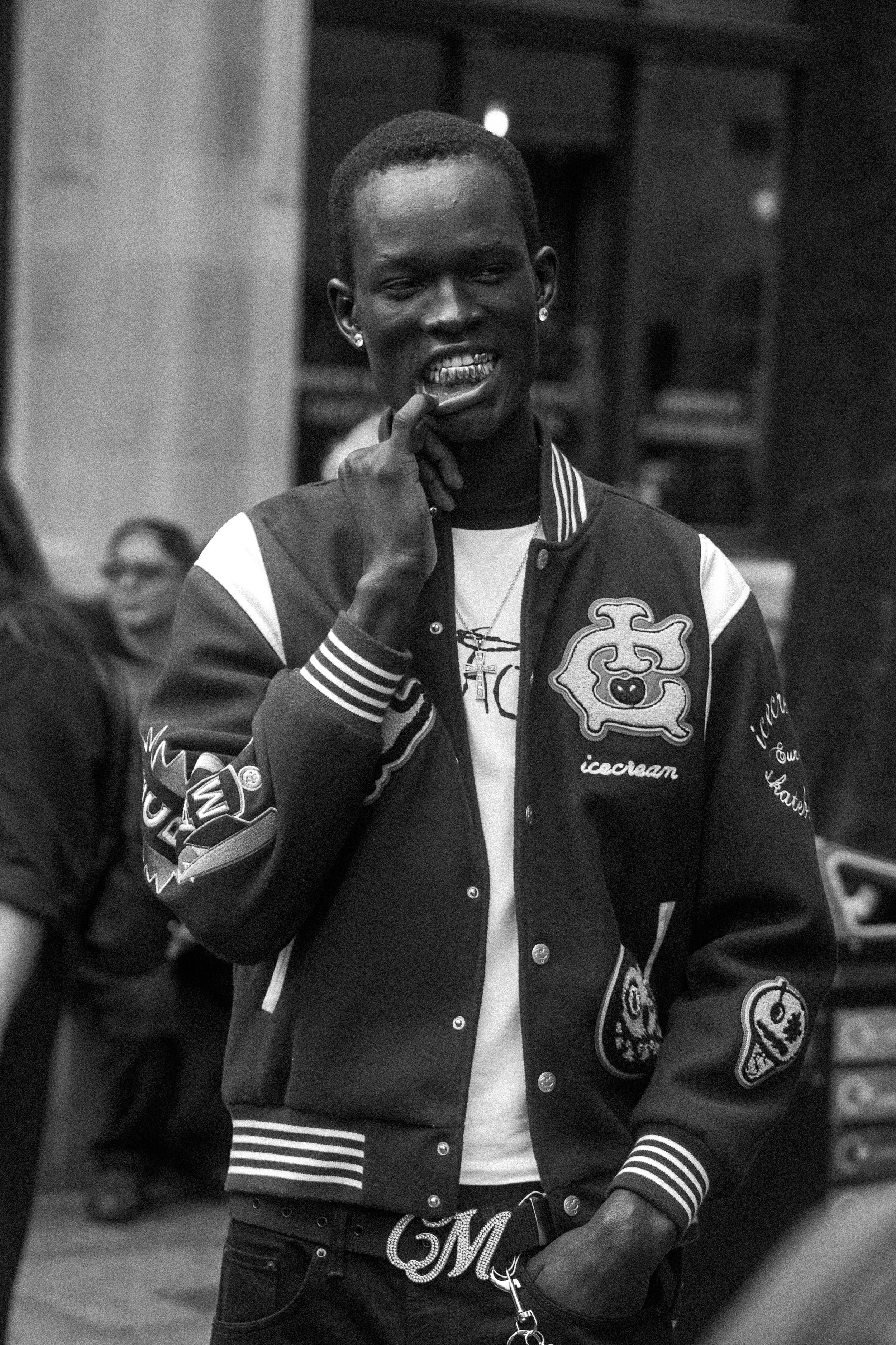A young man with dark skin and short hair, wearing a varsity jacket adorned with various patches, is seen biting his finger with a confident expression in an urban setting.