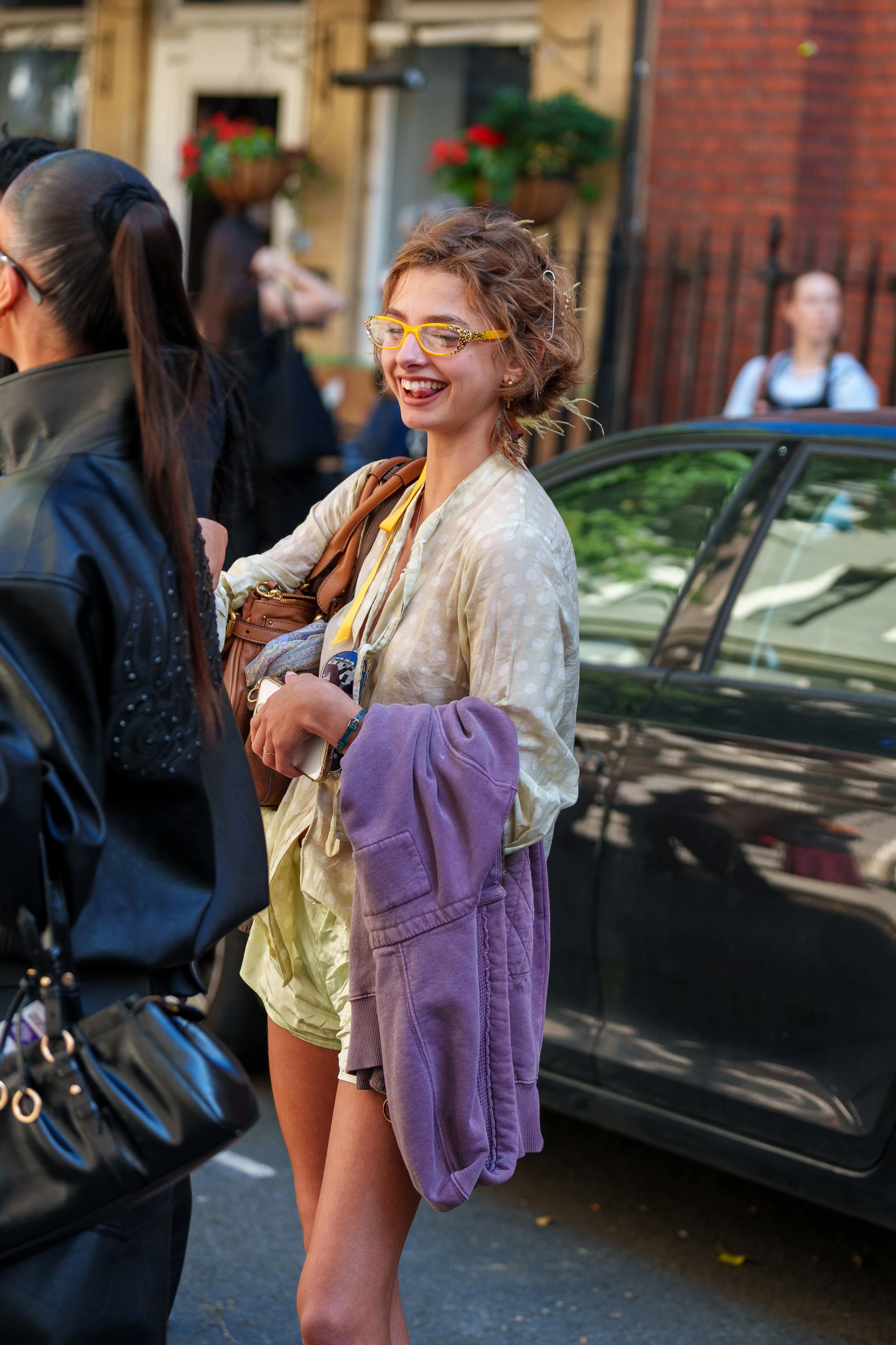 A smiling woman with curly hair and yellow glasses standing outdoors, holding a purple hoodie, with a brown bag over her shoulder, near a black car on a street with people and brick buildings in the background.