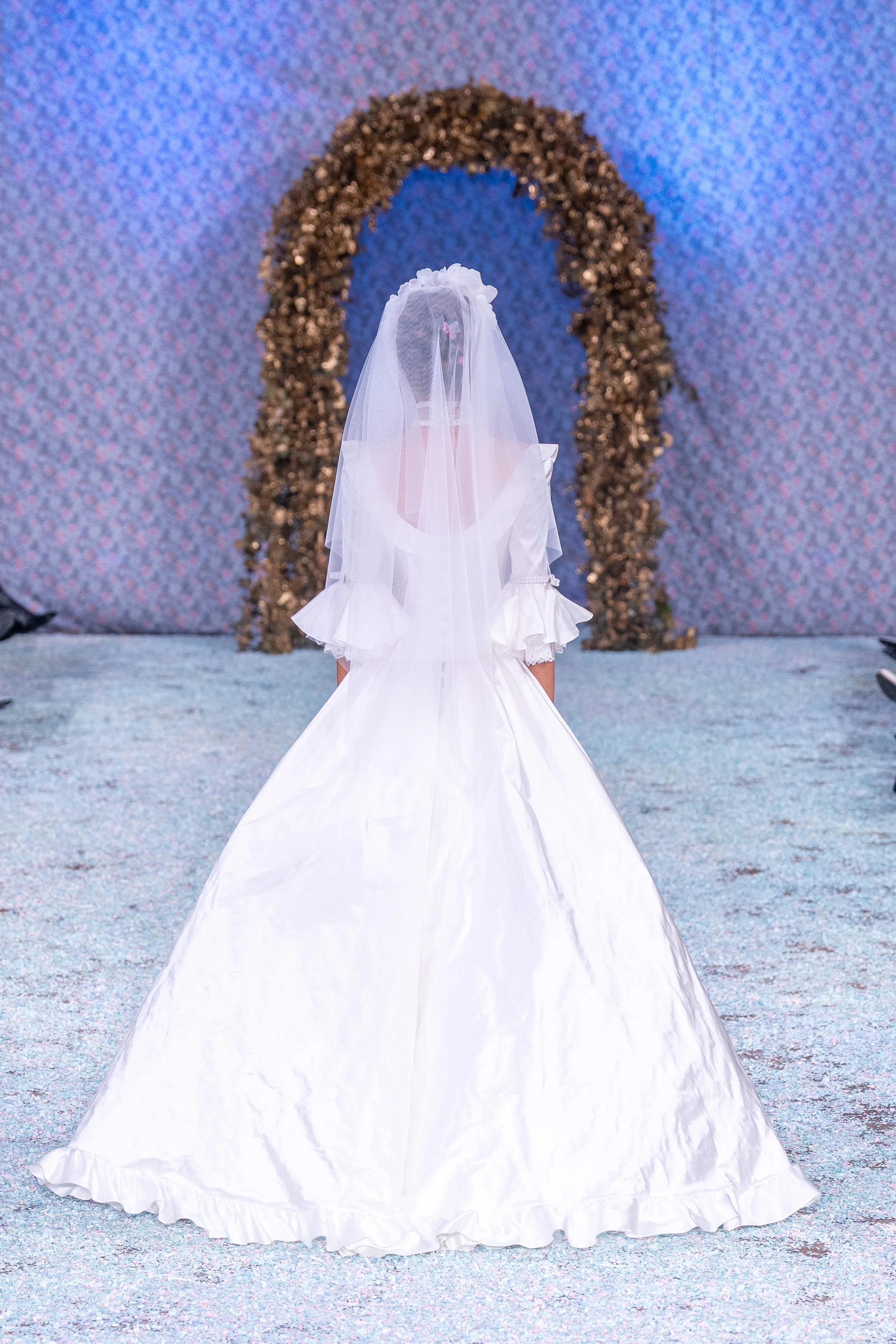 A young girl dressed in a white wedding gown with a veil is shown from behind, walking on a runway with a decorative arch in the background.