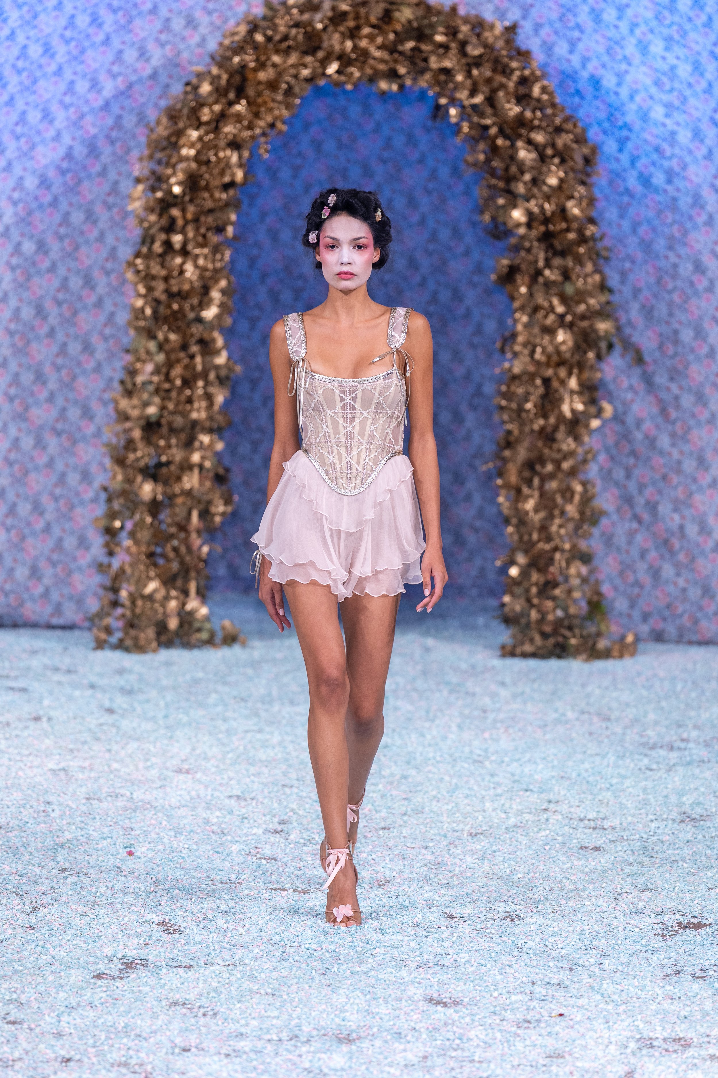 A model walking on a runway with a floral backdrop and gold arch, wearing a light pink, ruffled lingerie-style outfit with a corset top and matching heels decorated with flowers.