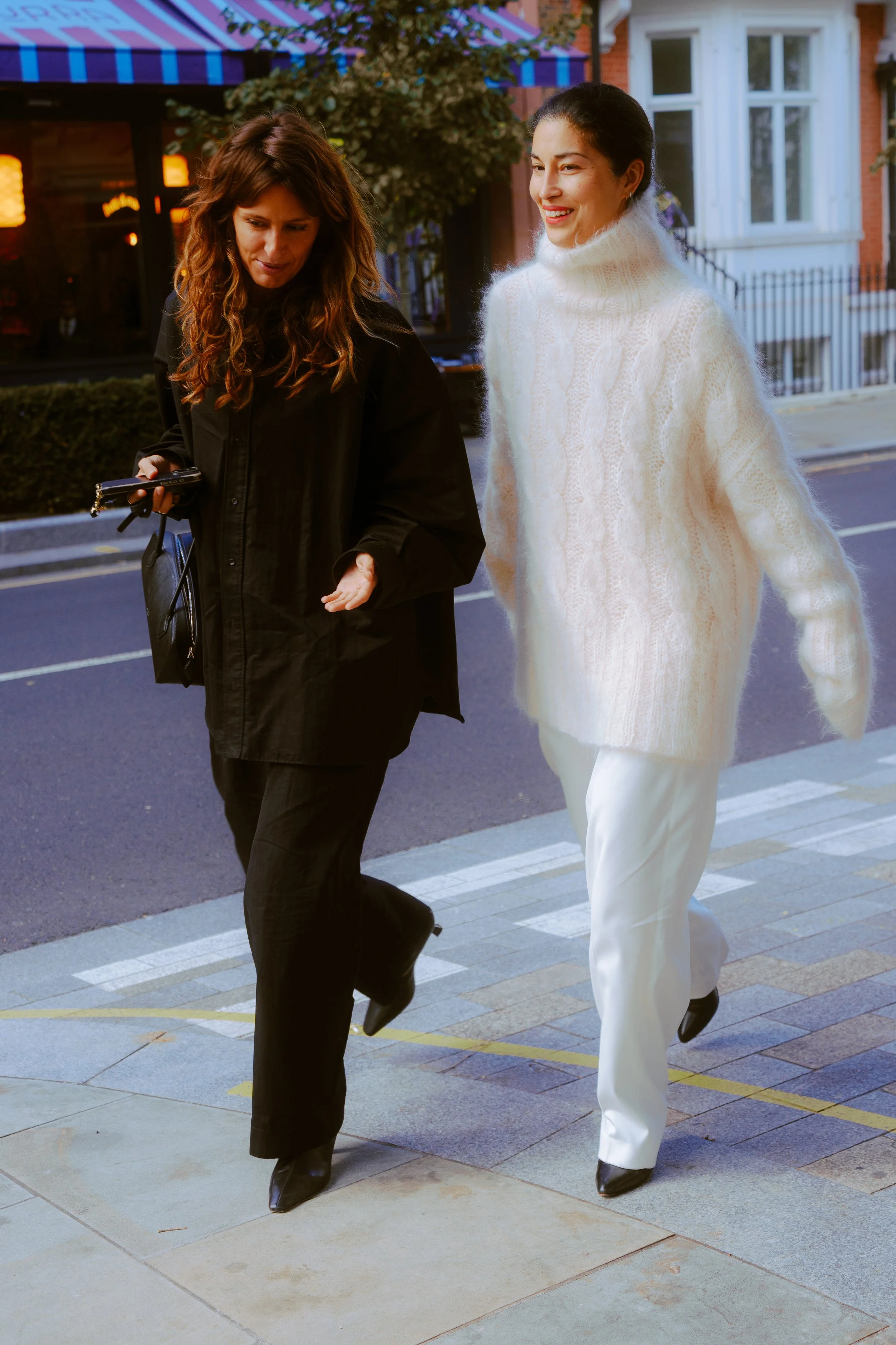 Two women walking on a city sidewalk, one wearing a black coat and pants, the other in a white turtleneck sweater and white pants, during evening time.