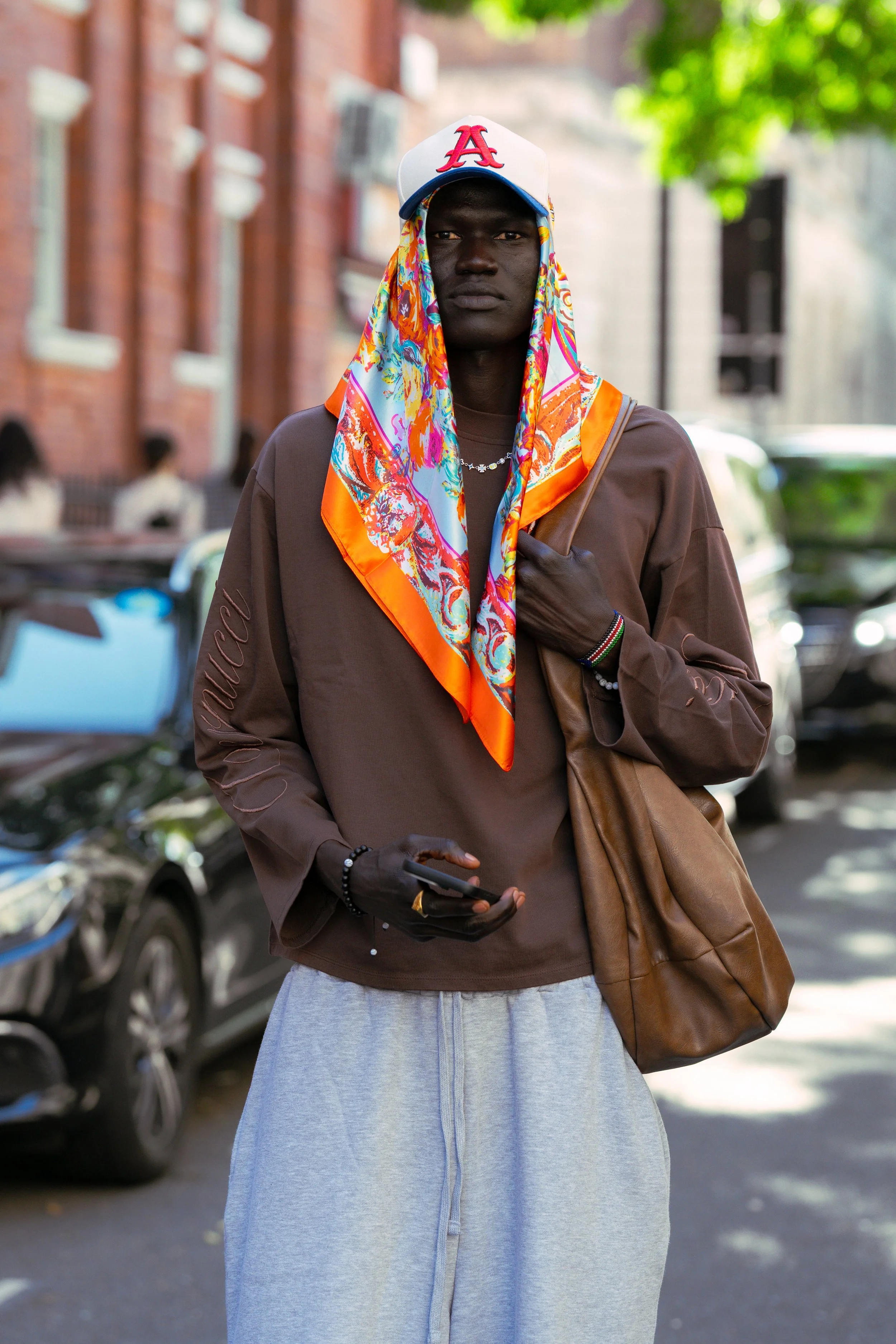 A young man with dark skin standing on a city street, wearing a white baseball cap with a red logo, a colorful scarf, a brown sweatshirt, and gray sweatpants. He holds a phone in his right hand and has a brown bag over his shoulder, with trees and ca