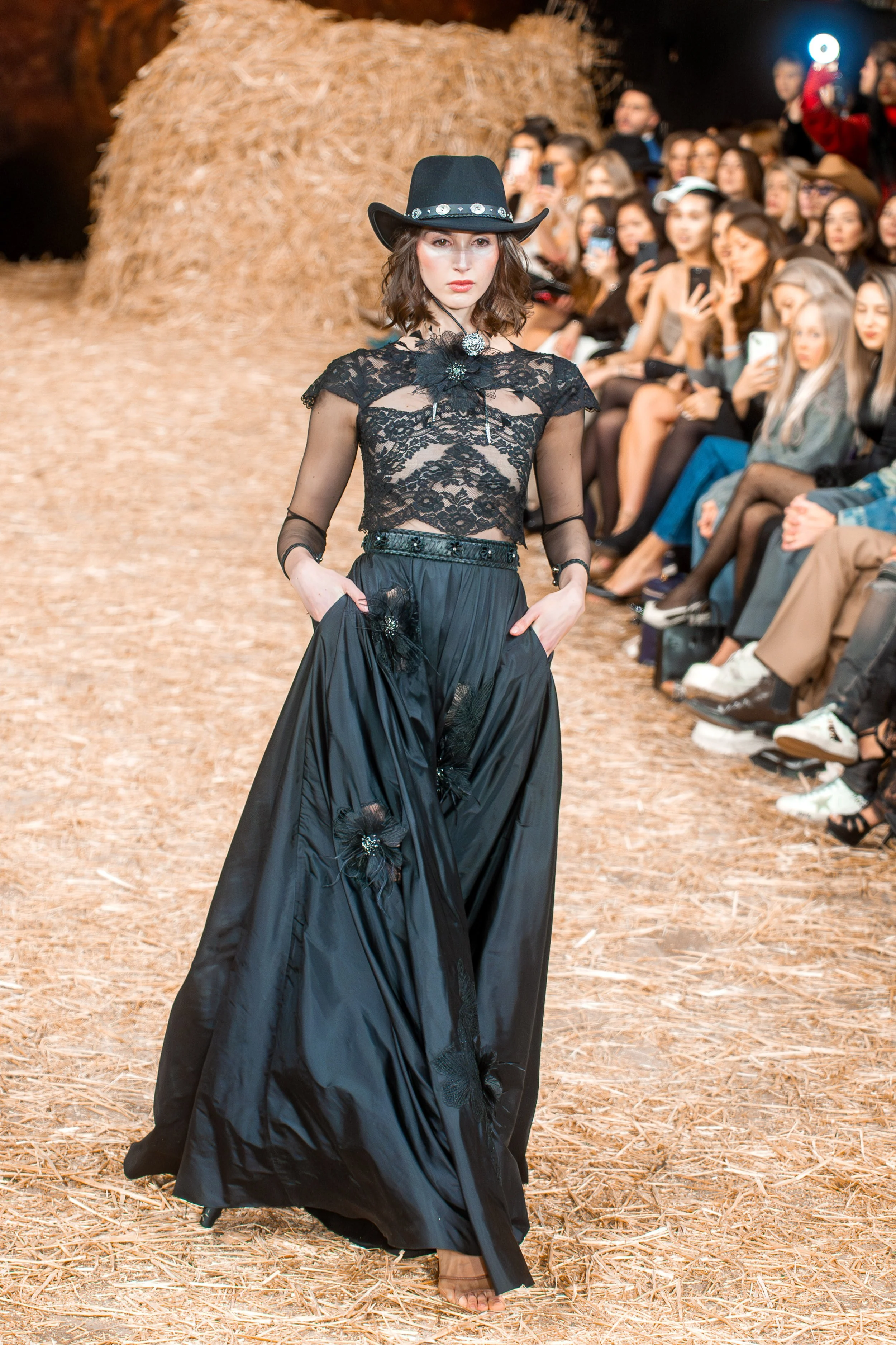 A female model walks down a runway wearing a black lace crop top with floral details and a long black satin skirt adorned with similar floral embellishments. She pairs the outfit with a black cowboy hat decorated with silver accents. The runway has a