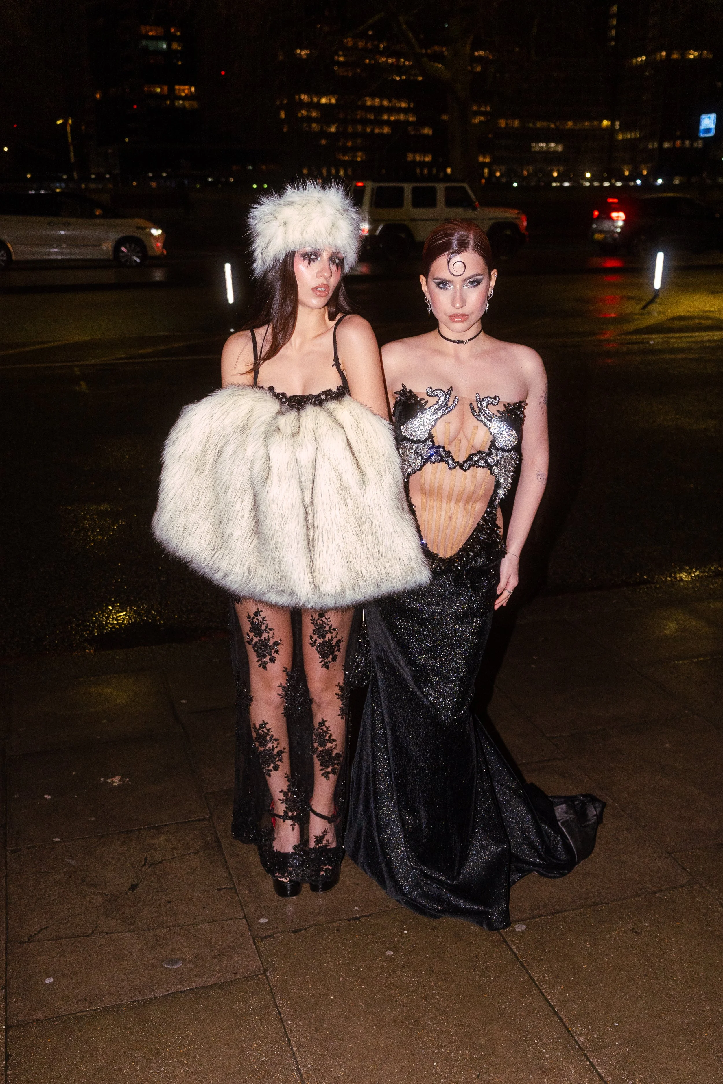 Two women in glamorous evening wear standing outdoors at night, with city lights and cars in the background.