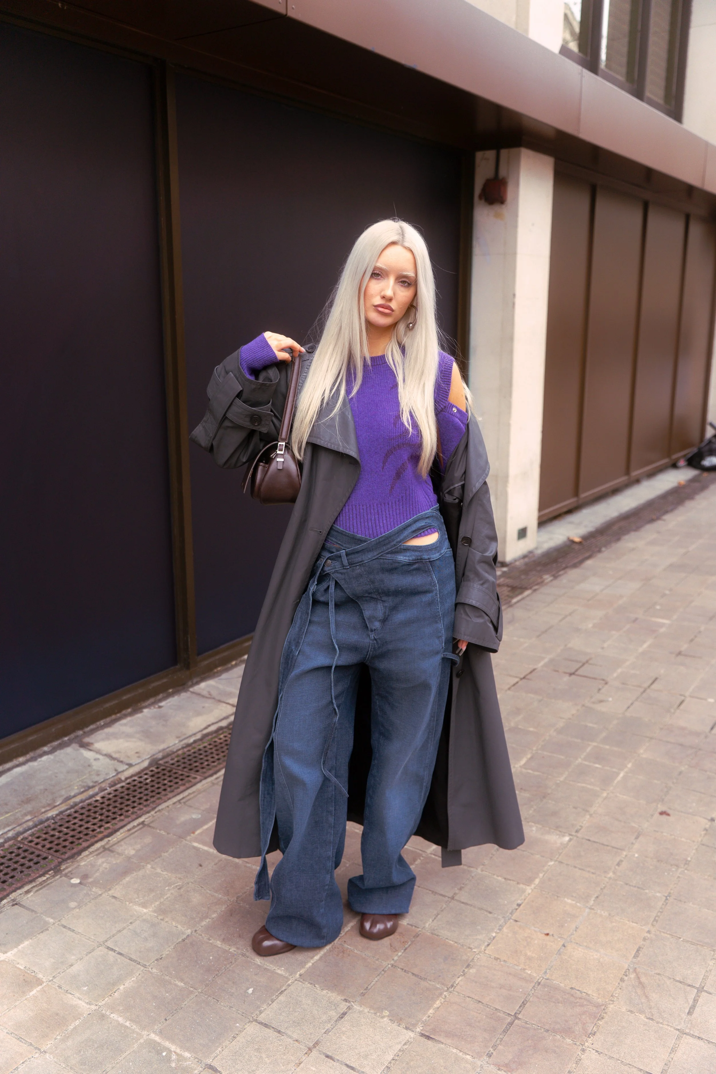 A woman with long platinum blonde hair standing on a city sidewalk, wearing a purple cut-out top, oversized dark blue jeans, a long gray coat, brown shoes, and carrying a small brown bag.