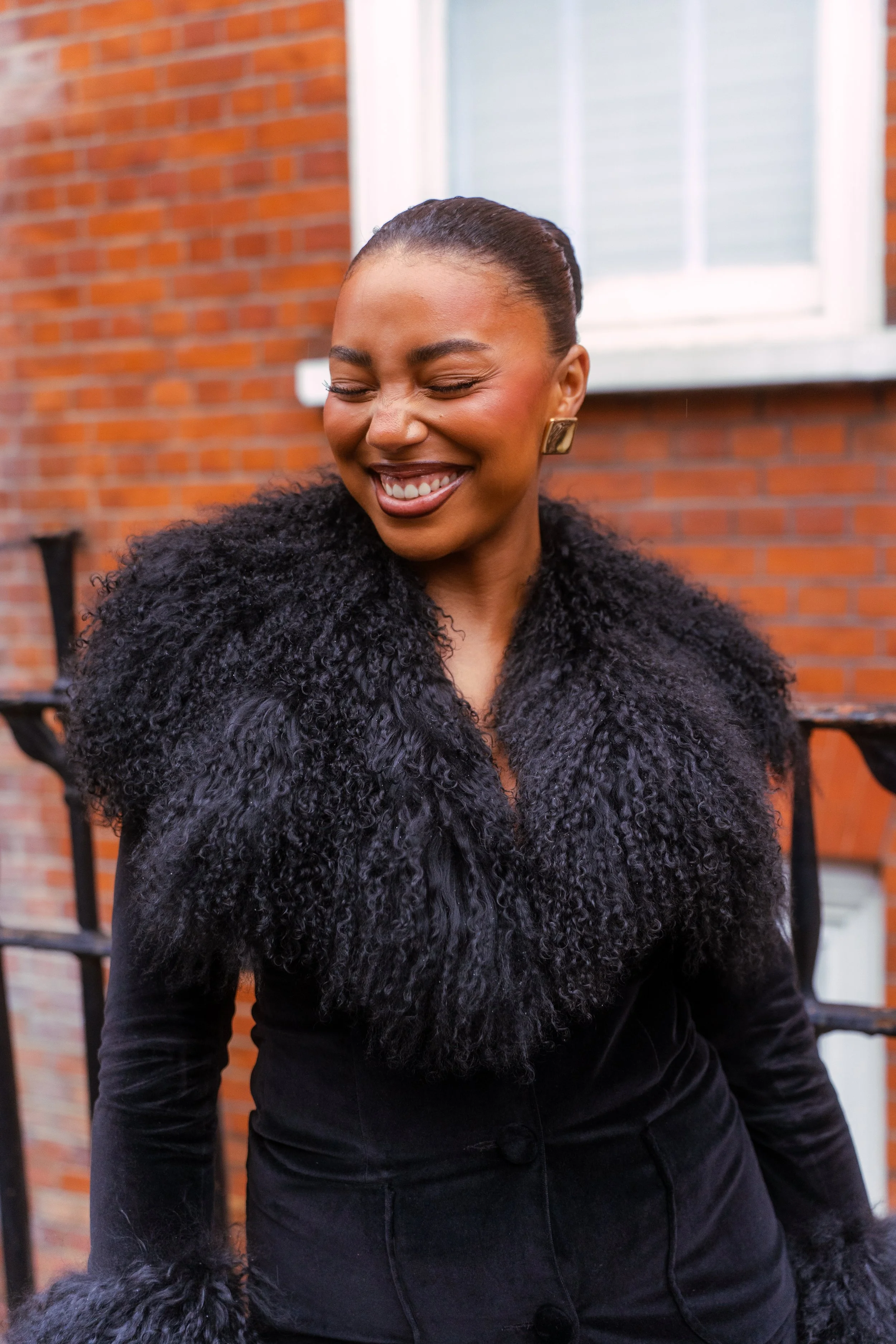 A woman with dark hair styled in an updo, smiling with her eyes closed, wearing a black faux fur coat, standing outdoors against a red brick building with a white window.