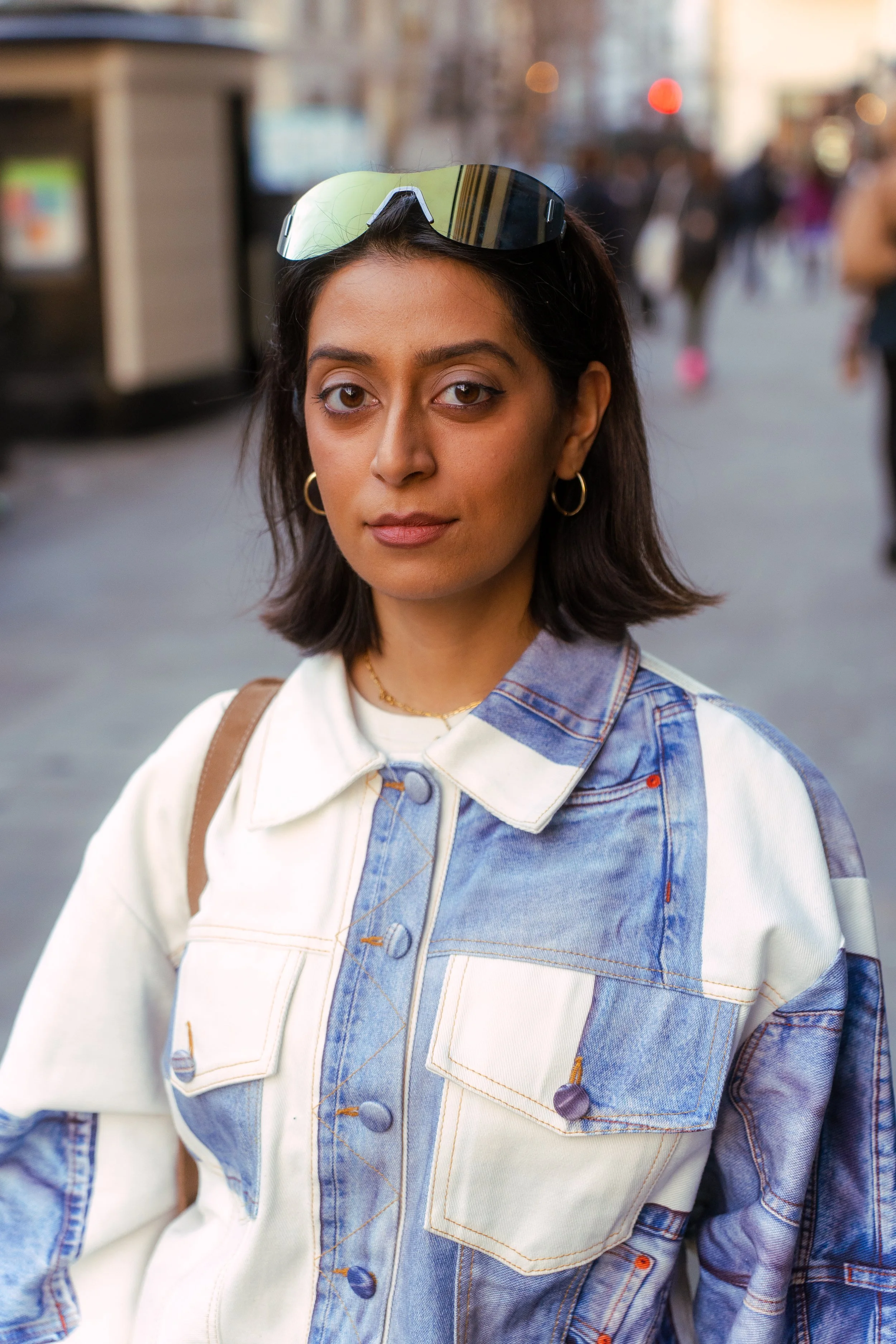 A young woman with shoulder-length dark hair wearing gold hoop earrings, a denim jacket with mixed color panels, and sunglasses on her head, standing on a busy city street with blurred pedestrians and buildings in the background.