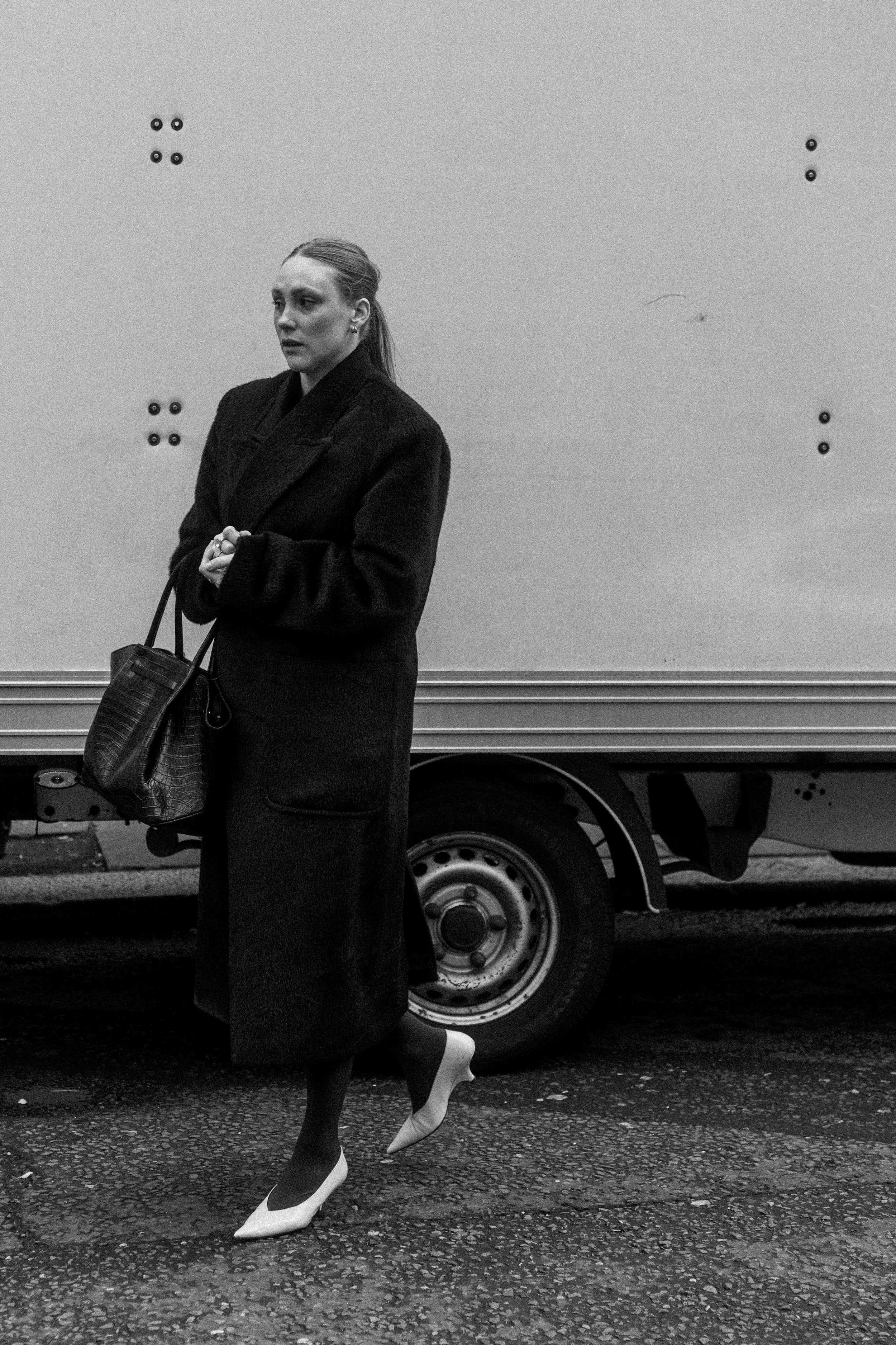 A woman in a long dark coat and high heels, holding a handbag, walking on a street with a moving truck in the background.