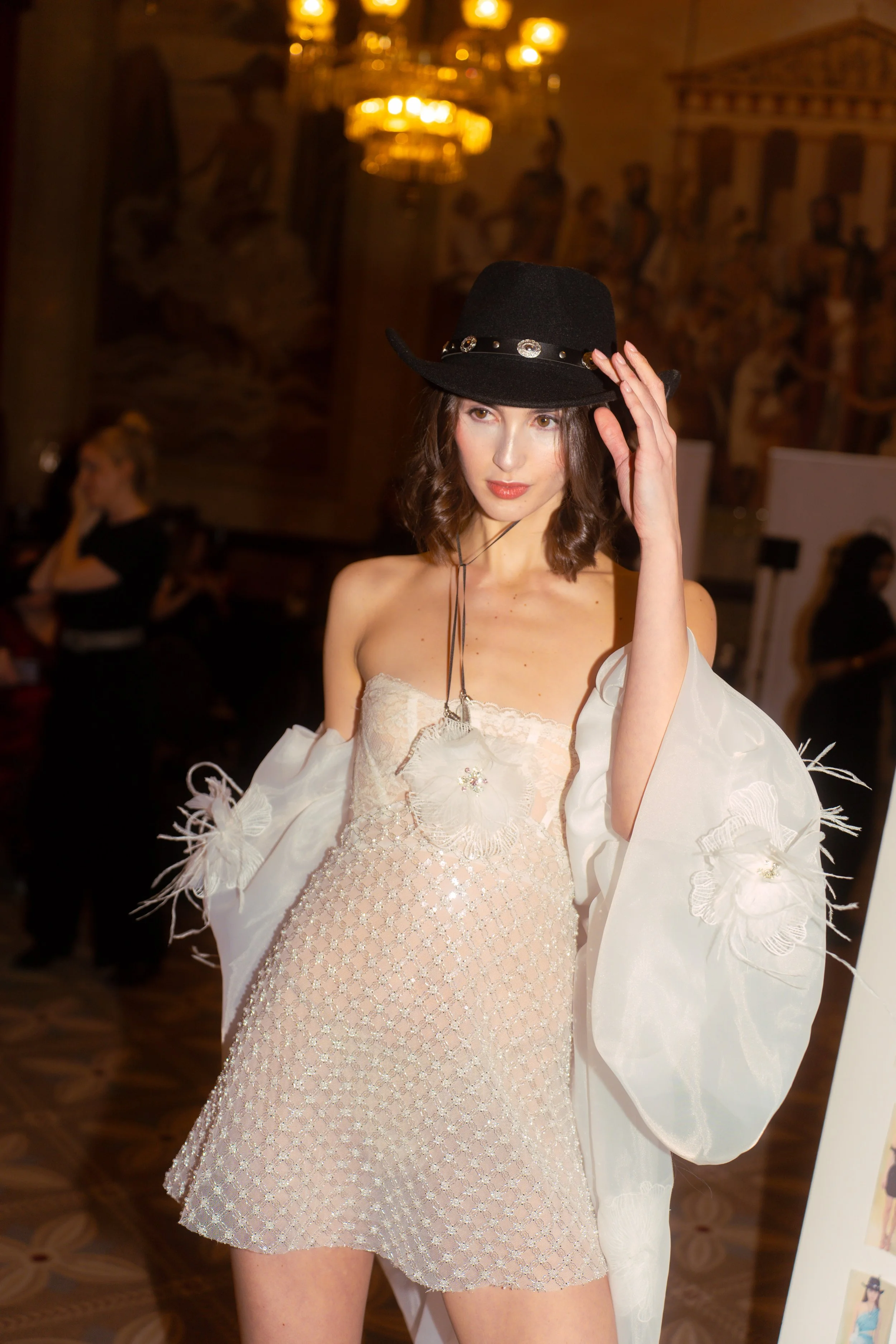 A woman with shoulder-length brown hair wearing a black hat, a strapless cream-colored dress with lace and embroidered details, and a white shawl with floral embroidery, standing indoors at a fashion event or gallery.