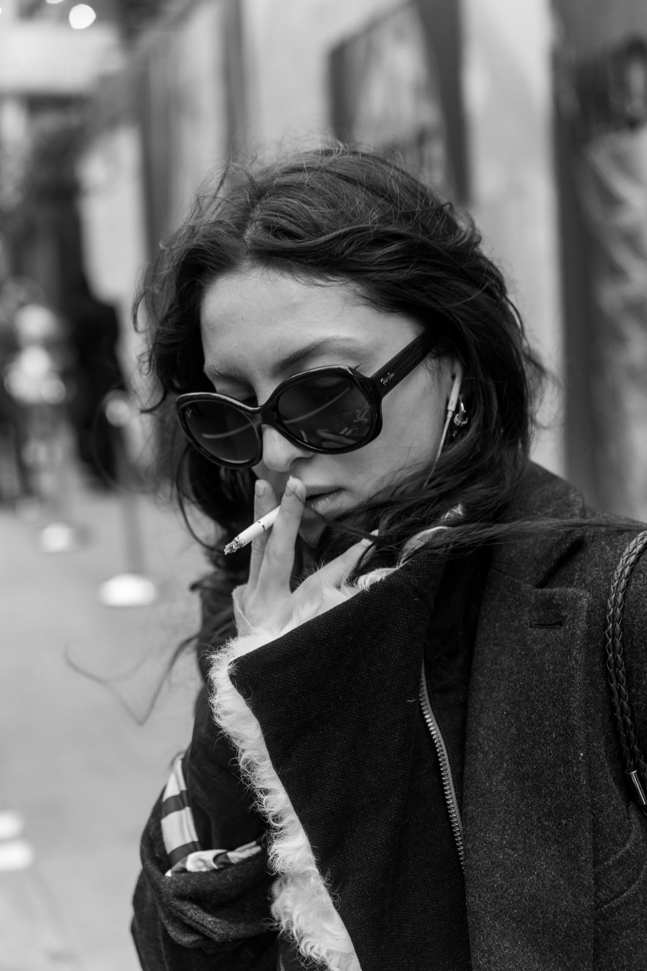 Black and white photo of a woman with wavy hair, wearing large sunglasses, holding a cigarette near her lips, and looking downward.