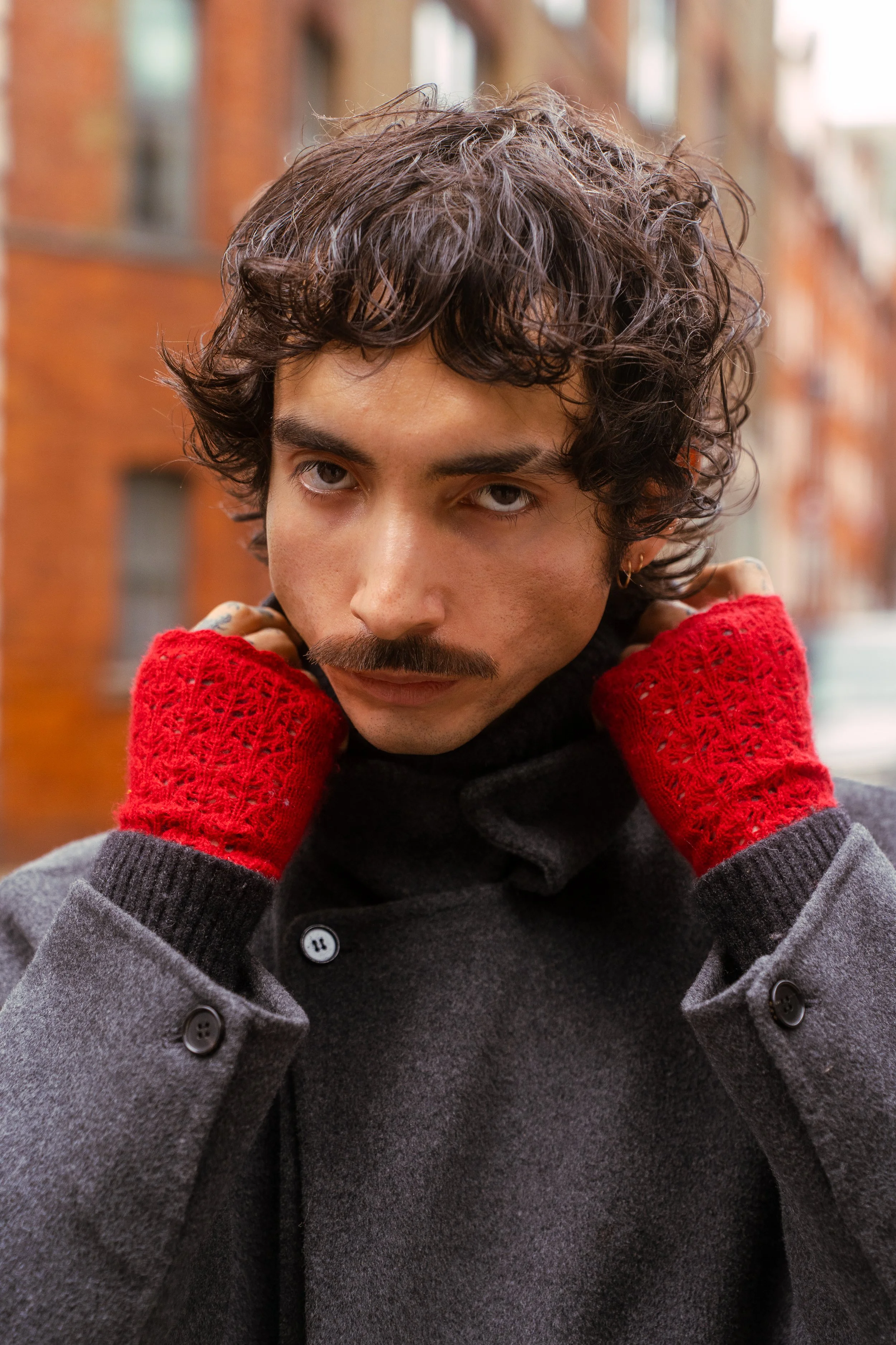 A young man with curly dark hair, a mustache, wearing a dark gray coat and bright red knit gloves, standing outdoors in front of a brick building, looking directly at the camera with a serious expression.