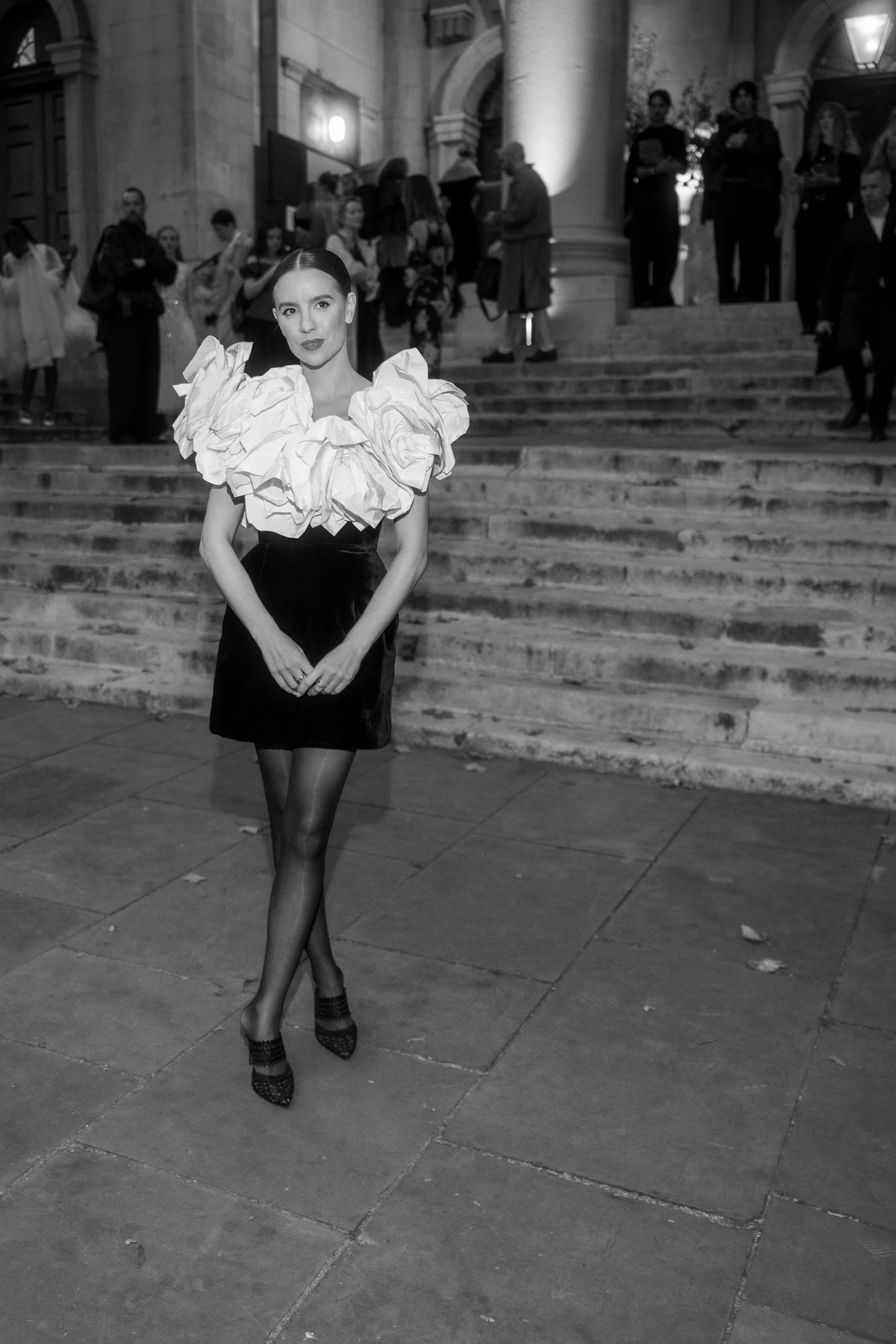 A woman stands in front of a staircase outside a building, dressed in a black dress with a large, ruffled white collar, black stockings, and high heels, during a nighttime event with other people in the background.