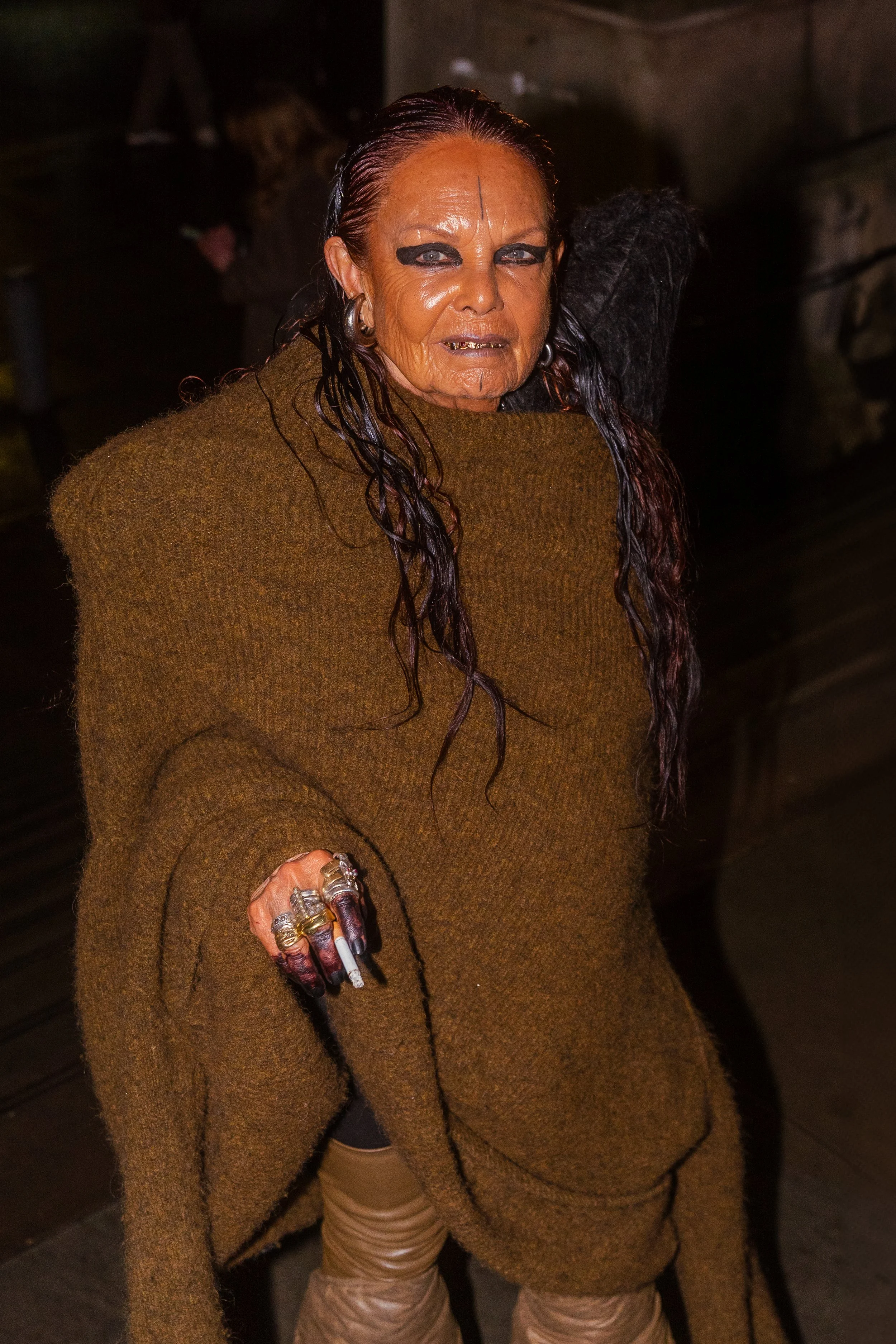 A woman with long wet hair, makeup, and multiple rings on her fingers, wearing a large brown sweater and tan pants, posing indoors at night.