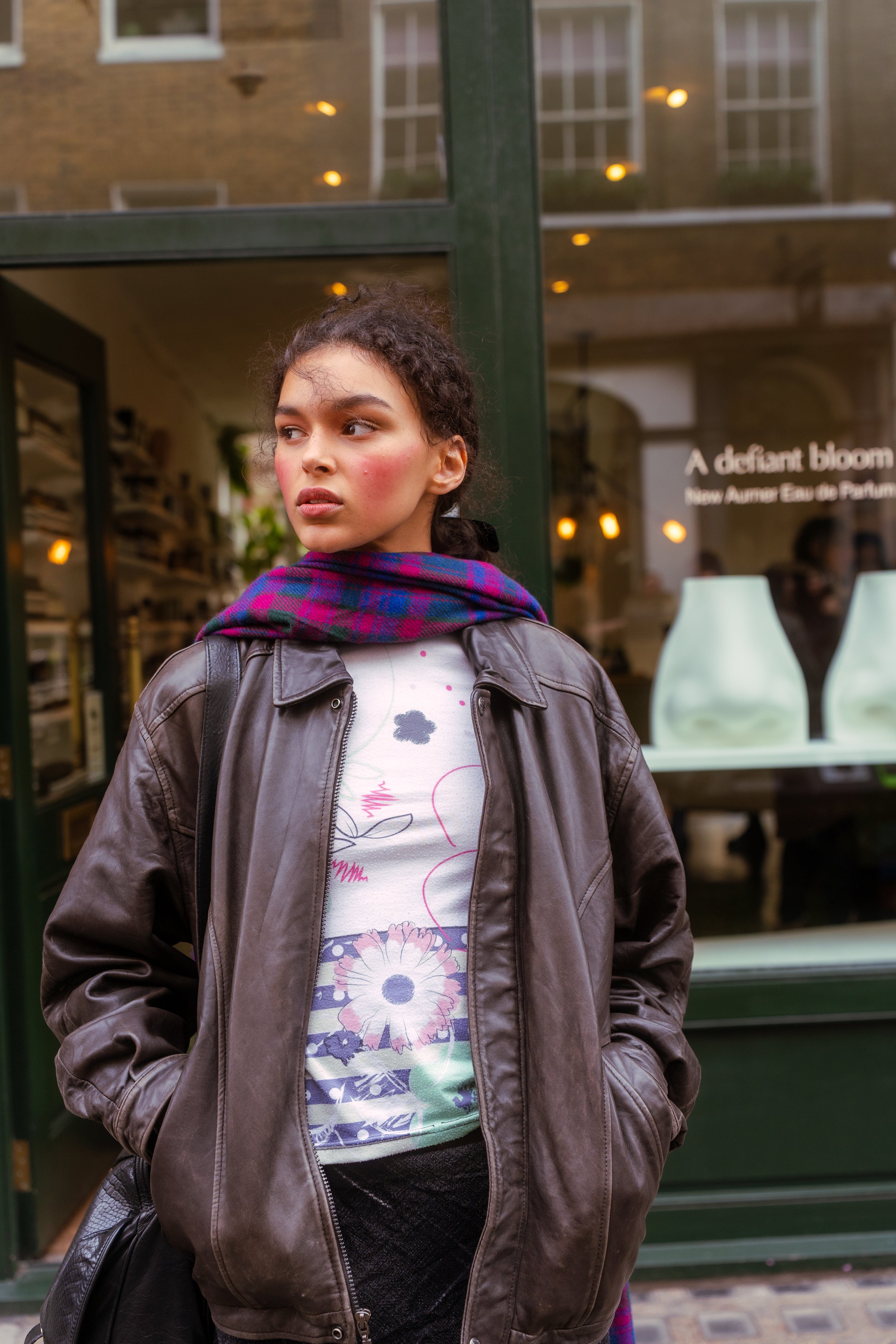A young woman with dark curly hair and light makeup, wearing a brown leather jacket, a colorful plaid scarf, and a floral shirt, stands outside a shop window with her hands in her pockets, looking to the side.