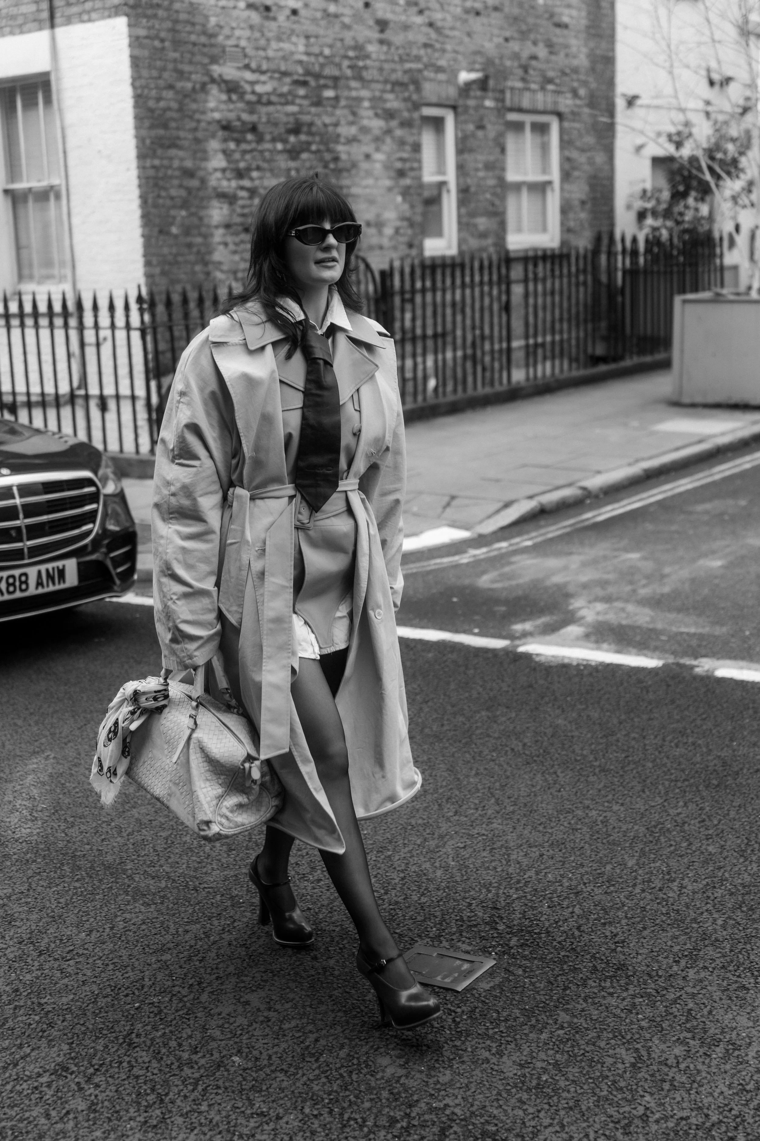 Black and white photo of a woman walking on a city street, wearing sunglasses, a trench coat, high heels, and carrying a handbag.