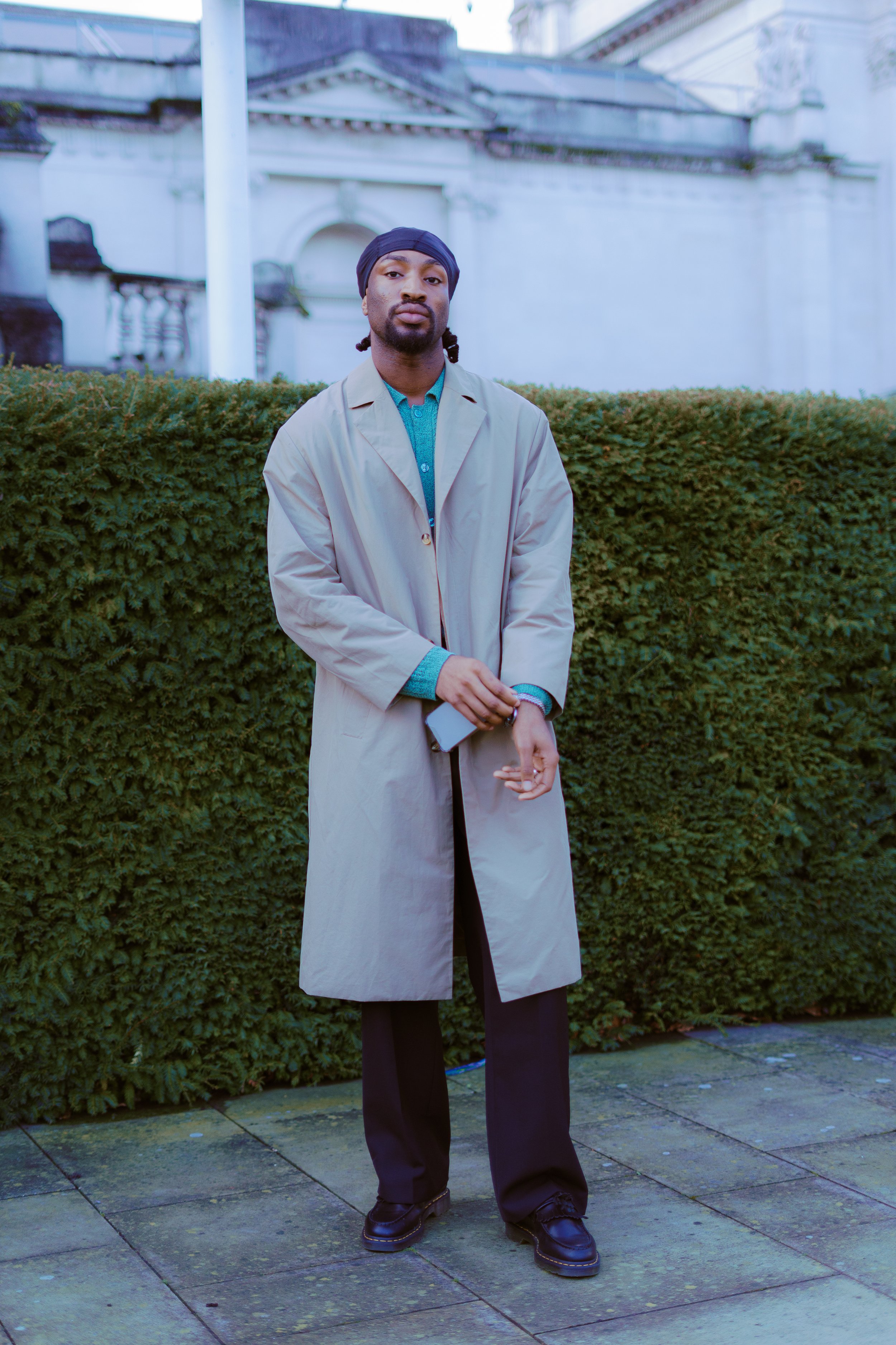 A man standing outdoors in front of a neatly trimmed hedge, wearing a beige trench coat, black pants, black shoes, and a navy blue headscarf, with a serious expression.
