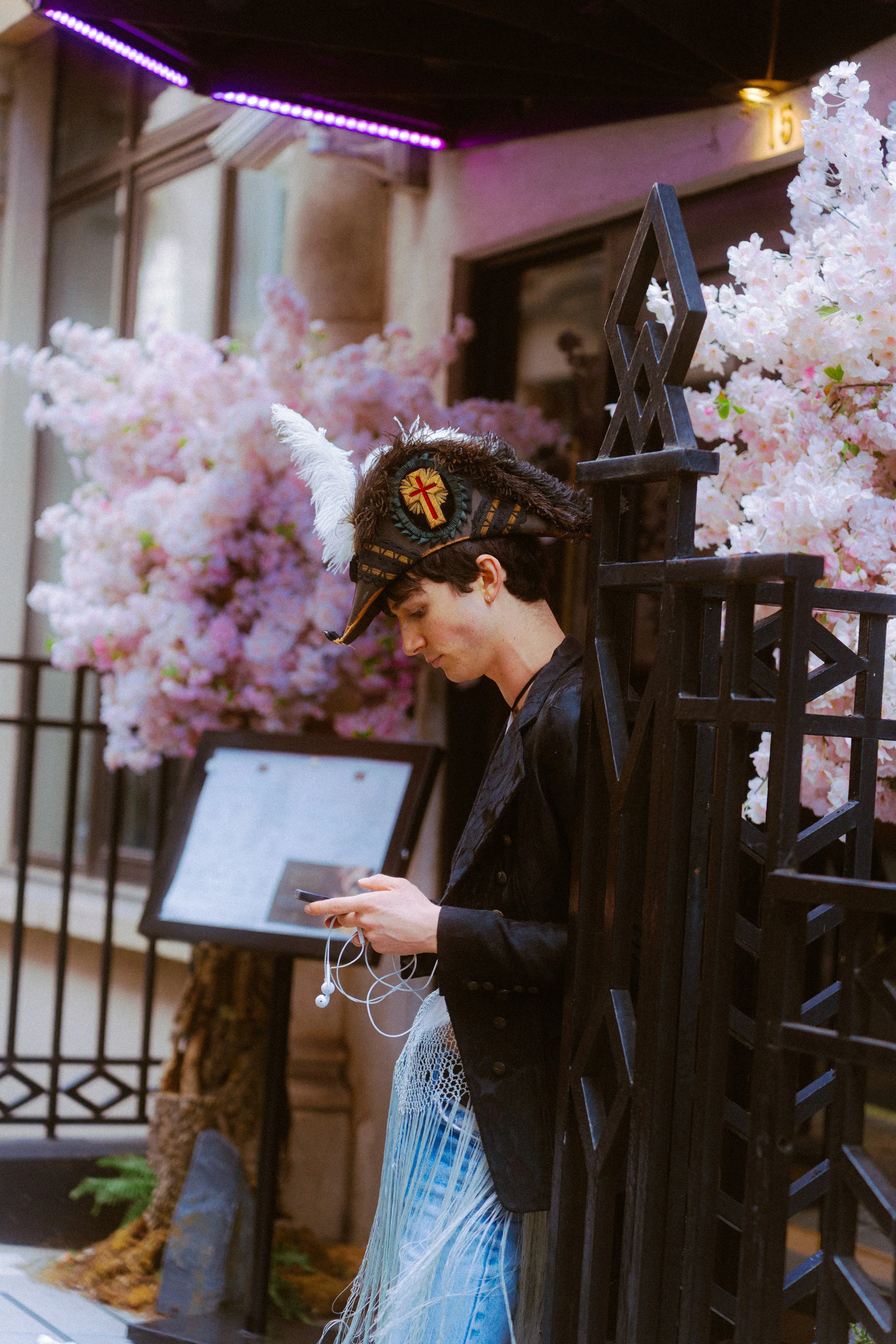 Young man wearing a Viking-style helmet with a feather, using his phone near pink cherry blossom trees and a black metal gate.