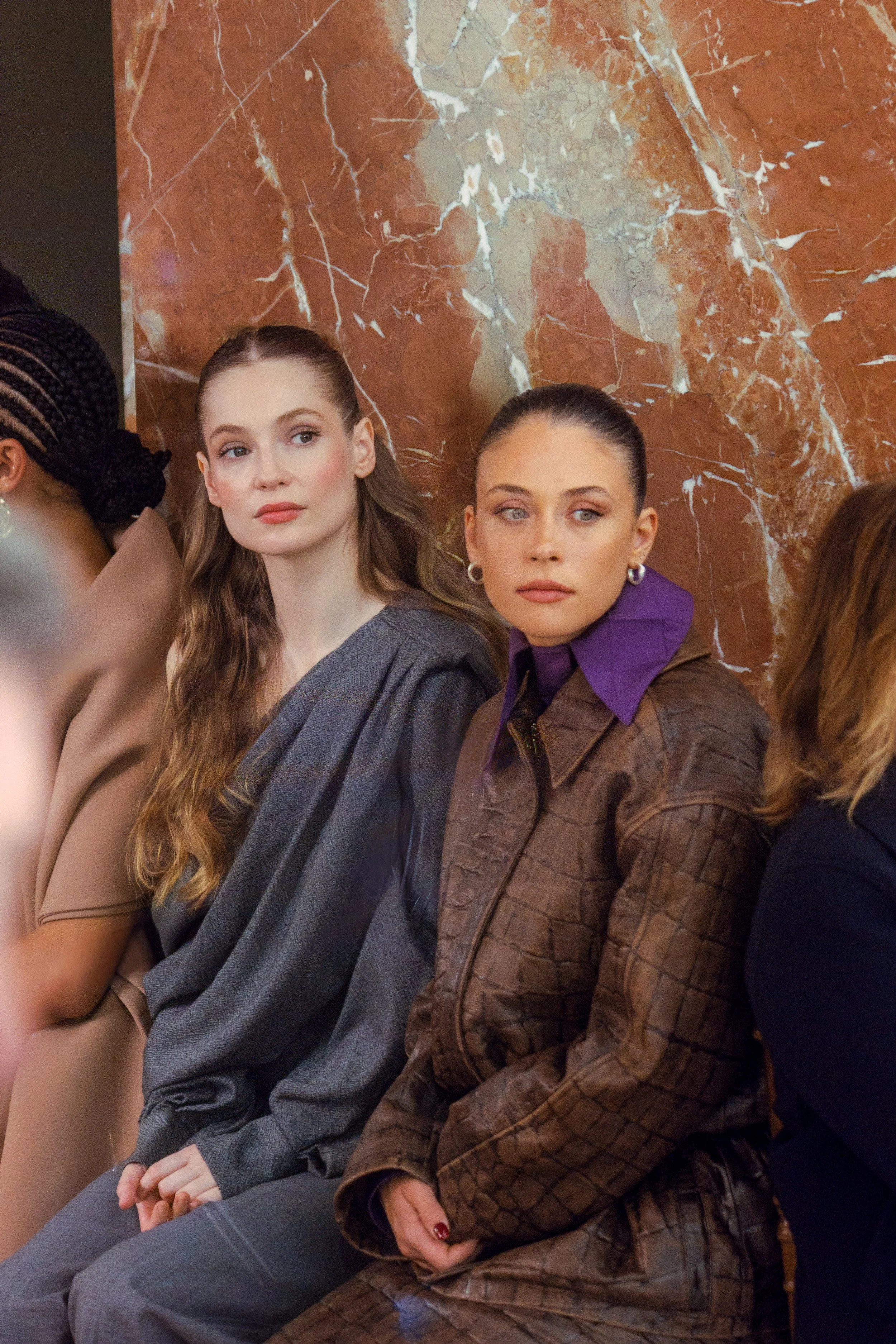 Two women sitting next to each other, one with long wavy hair wearing a gray blazer, and the other with dark hair in a bun wearing a brown leather jacket with a purple collar, on a marble wall background.