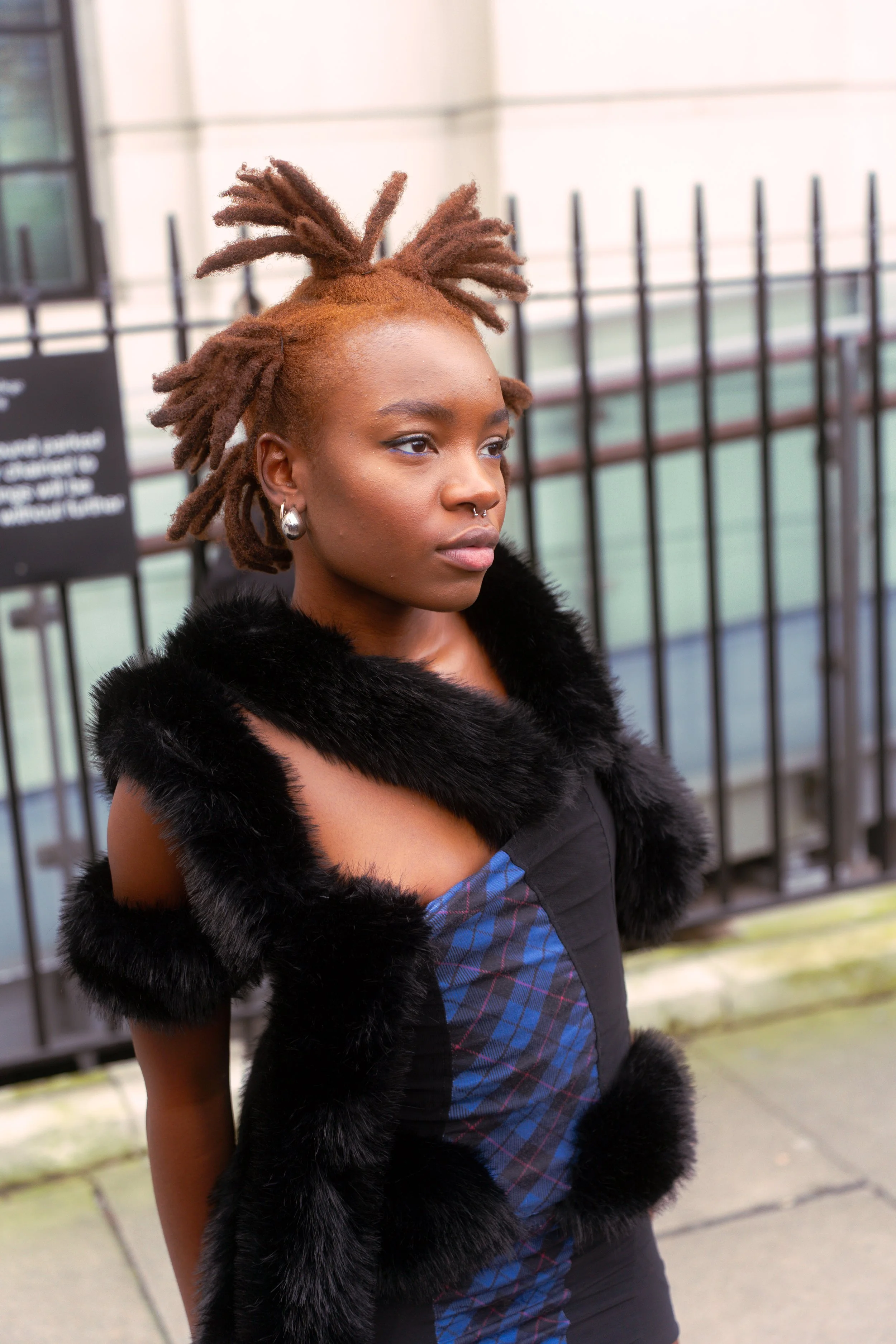 A young woman with reddish-brown dreadlocks, wearing earrings, a black fur-trimmed dress, and standing outdoors by a fence.