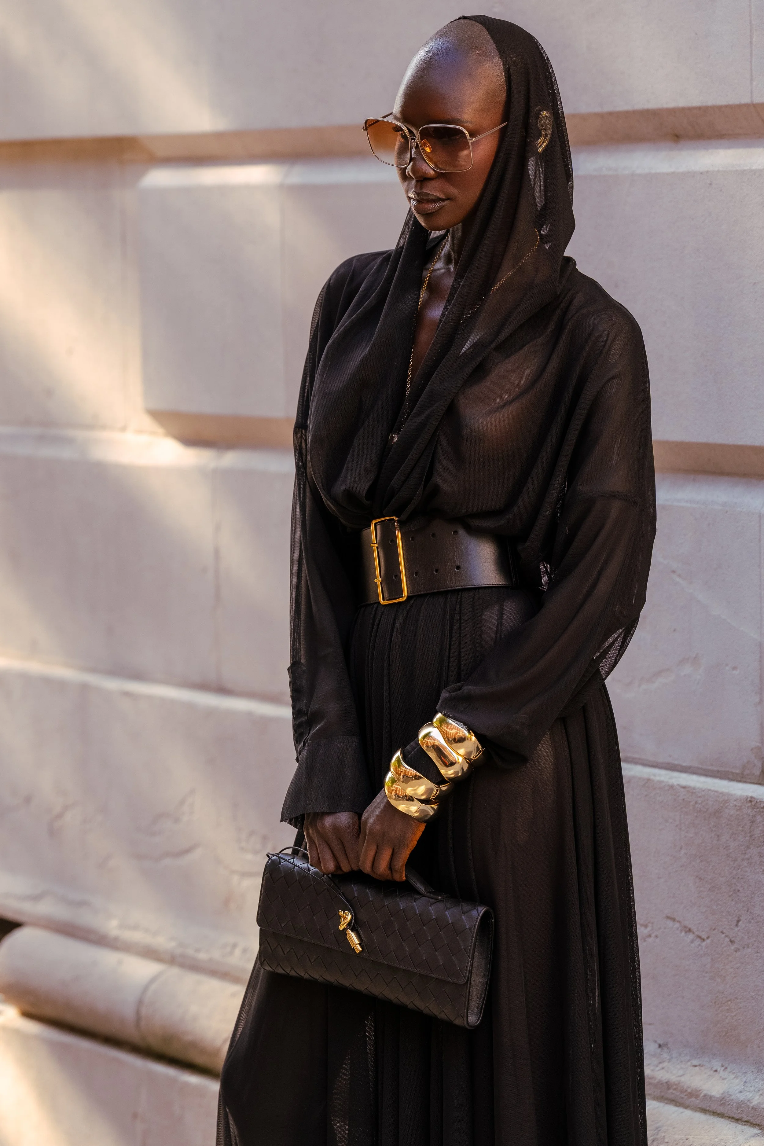 A woman dressed in black, wearing sunglasses, gold jewelry, and holding a black quilted handbag, standing against a light-colored stone wall.