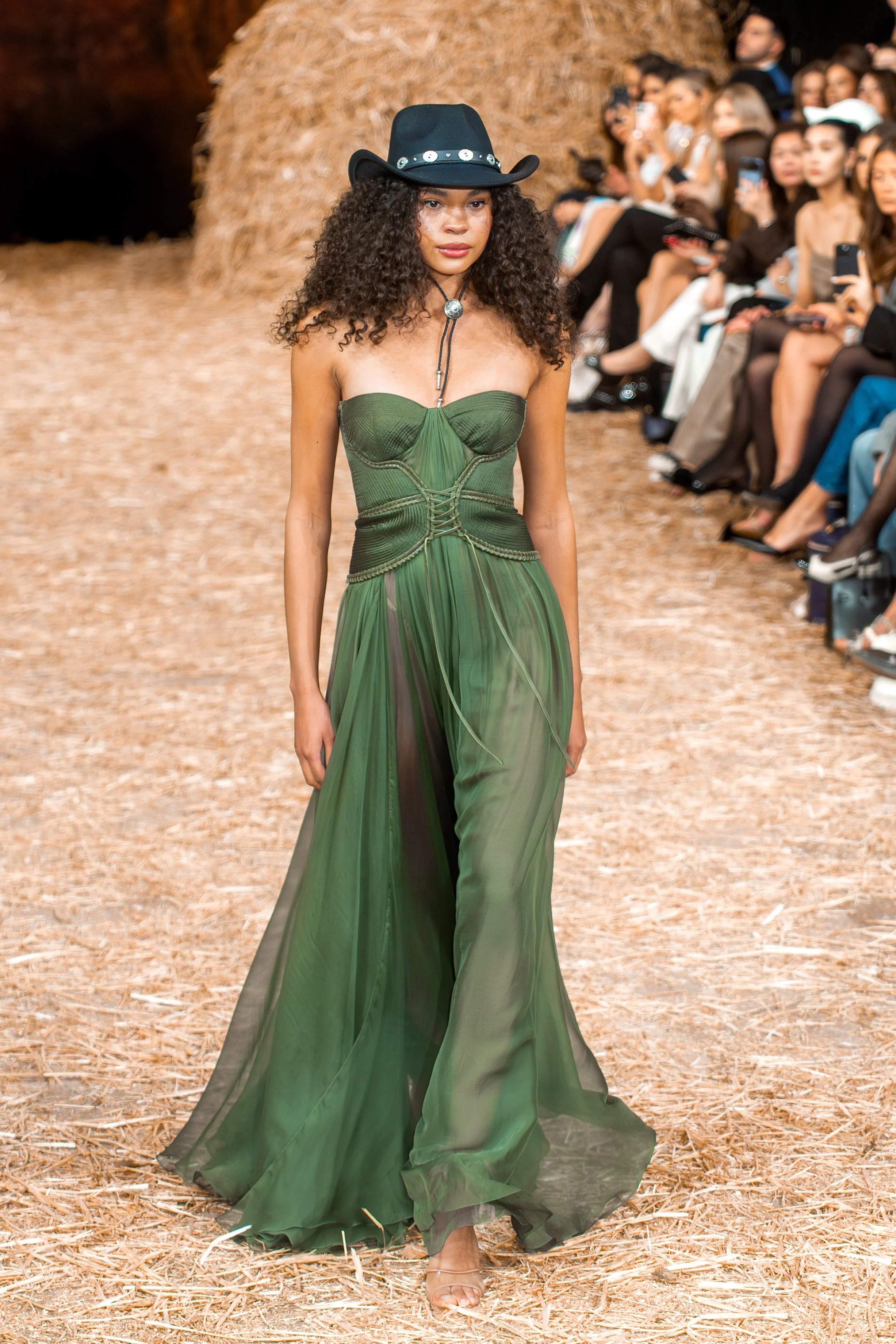 Model walking on a runway in a green, strapless, chiffon dress with a black cowboy hat, during a fashion show attended by an audience seated on both sides.