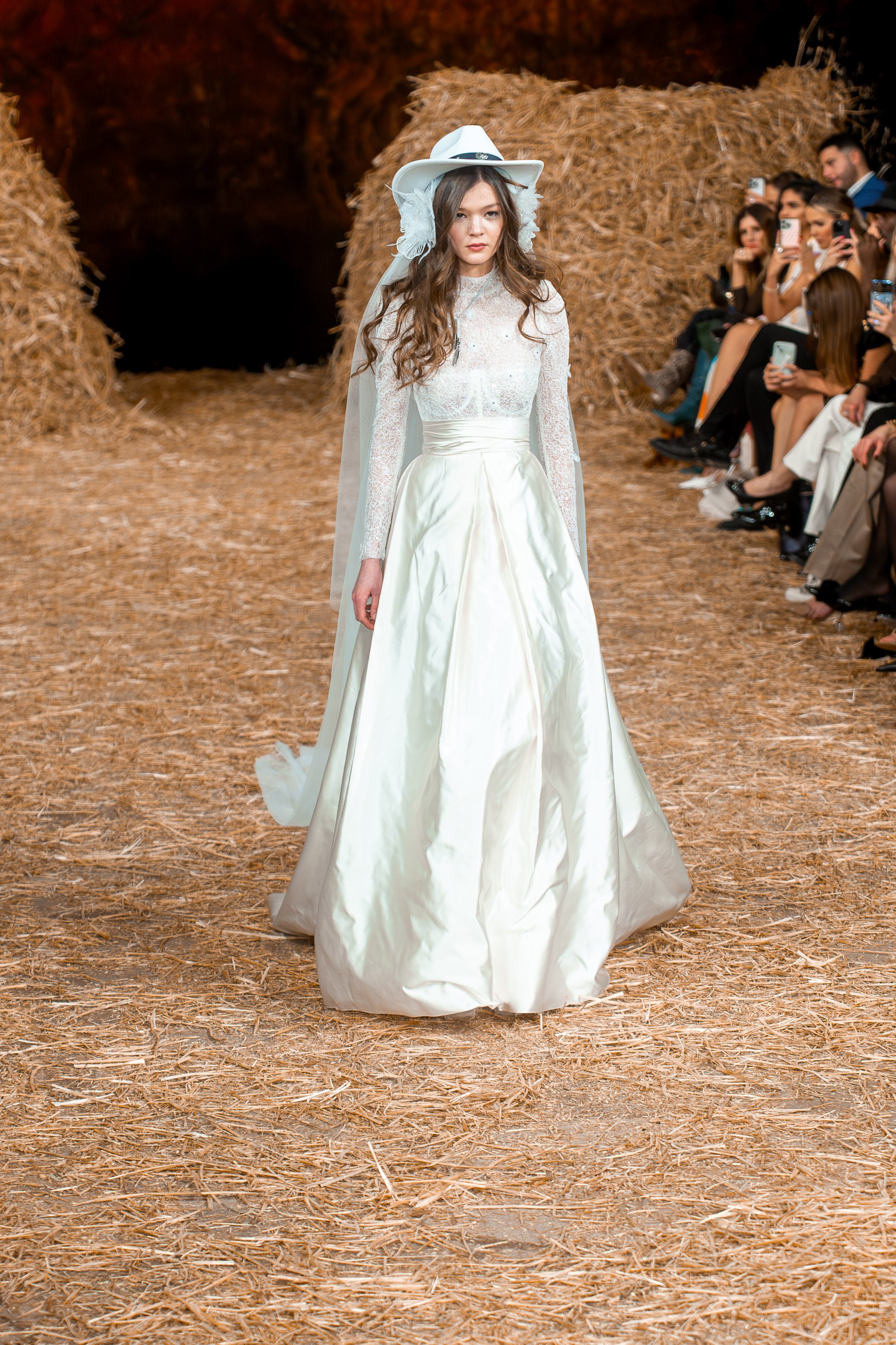 A female model walking down a runway in a wedding dress, with a crowd of spectators seated on her right side, and hay bales and a dark background behind her.