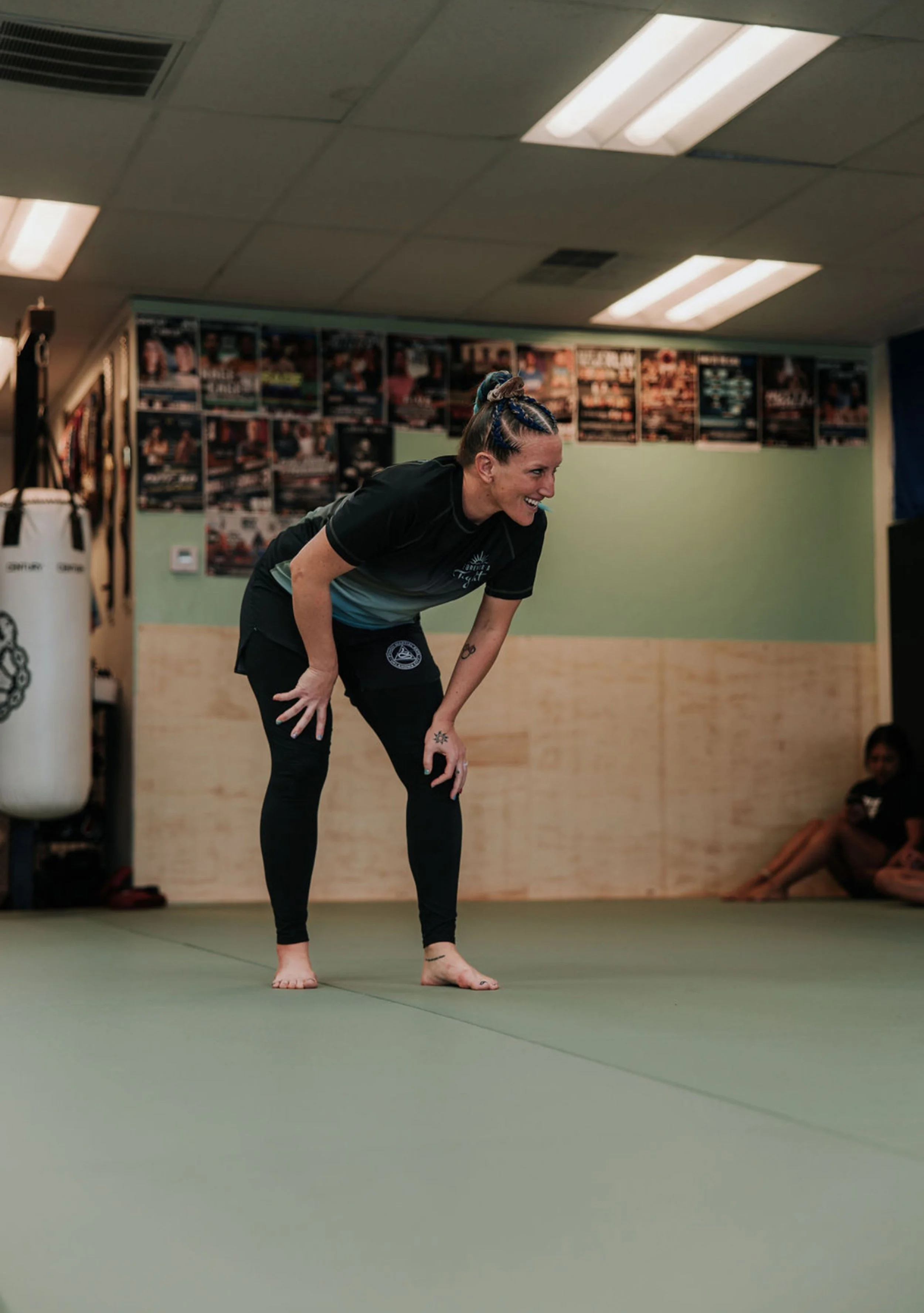 A woman in athletic wear bending forward in a martial arts or fitness training studio, smiling. She has braided hair with blue highlights, and people sitting against the wall in the background.