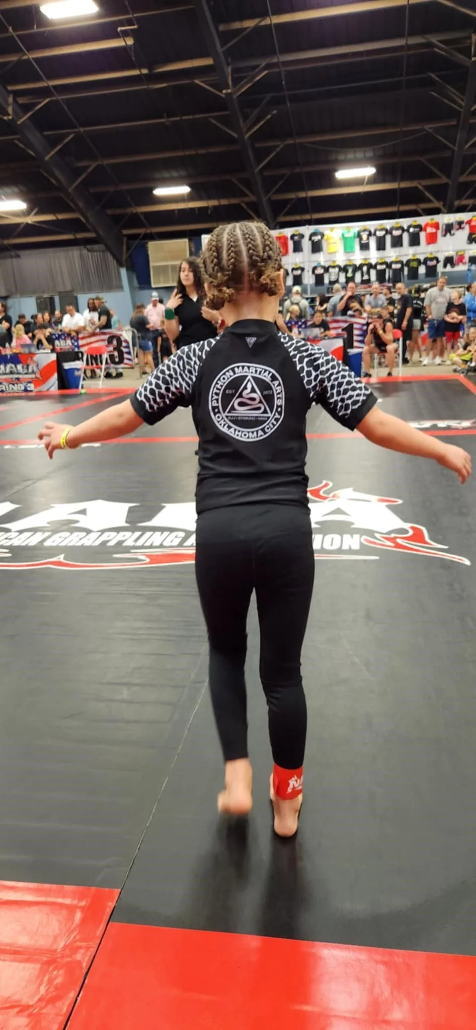 A young girl with braided hair standing on a martial arts mat at a grappling competition, wearing a black uniform with the logo of Pytion Martial Arts Oklahoma City on the back, surrounded by spectators and competitors.