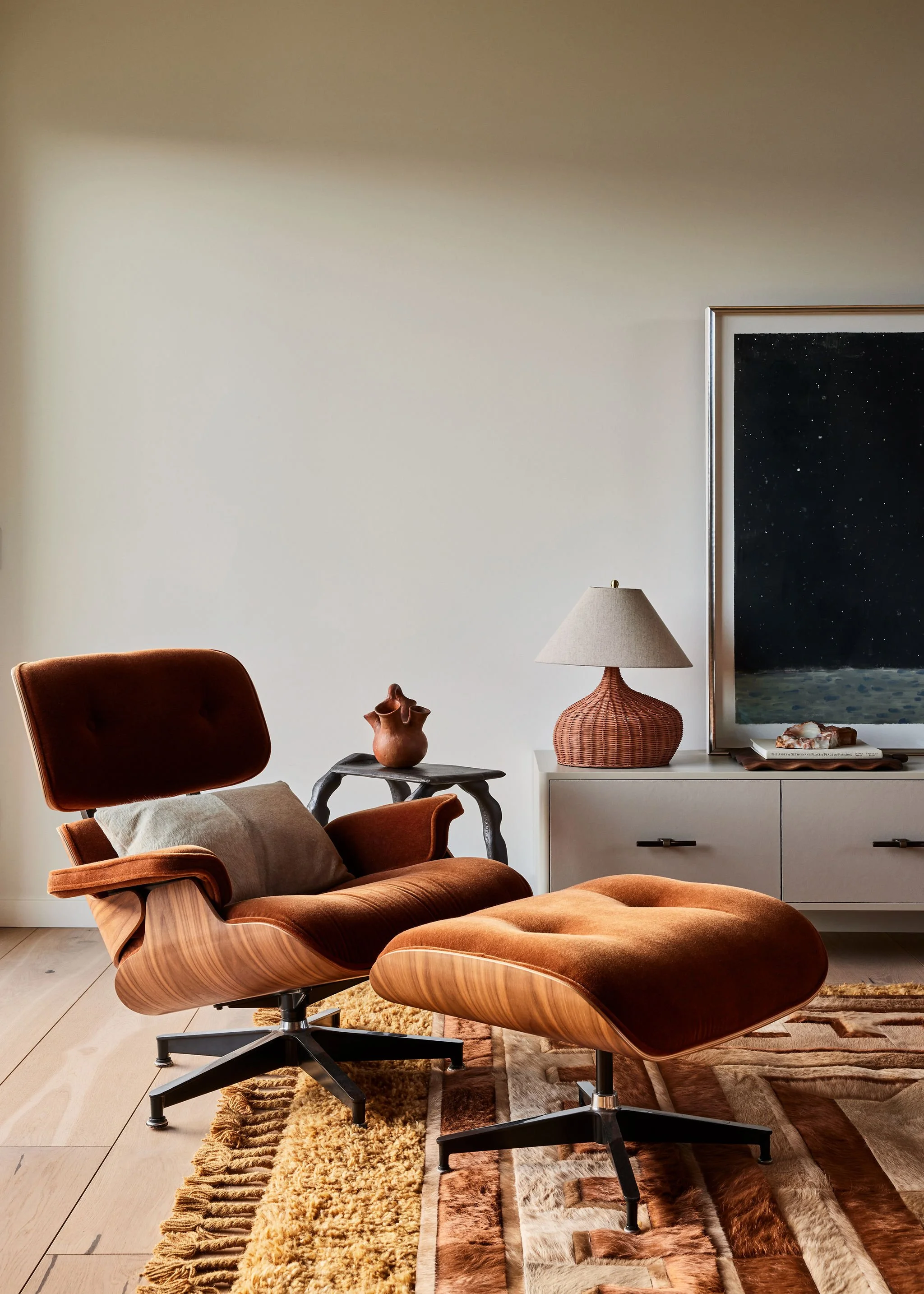 Living room with a brown leather armchair with matching footrest, a side table with a decorative vase, a white sideboard with a lamp and framed artwork, and a patterned rug on light wood flooring.