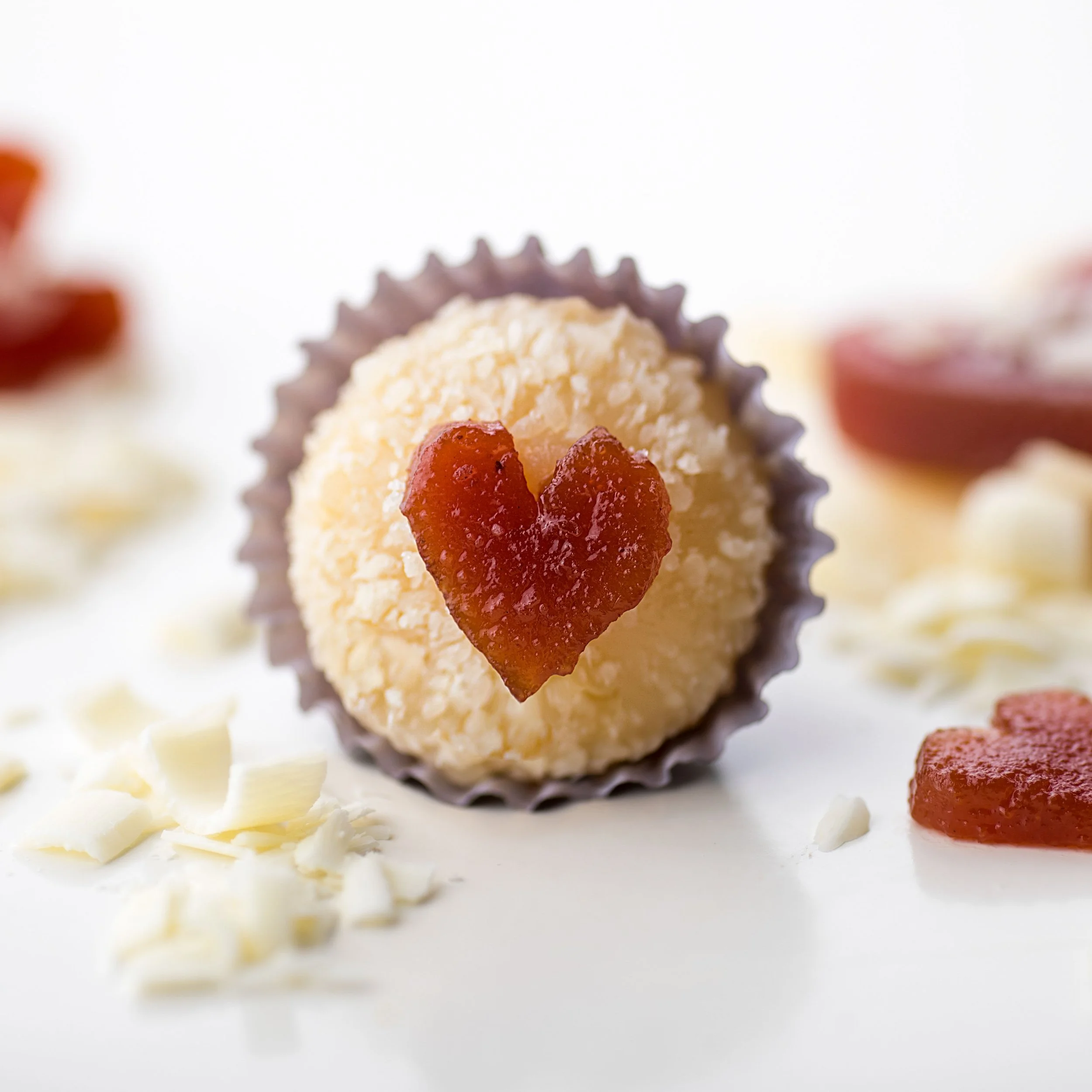 A coconut truffle with a heart-shaped red topping on a white surface, surrounded by white chocolate shavings and another red heart-shaped piece.
