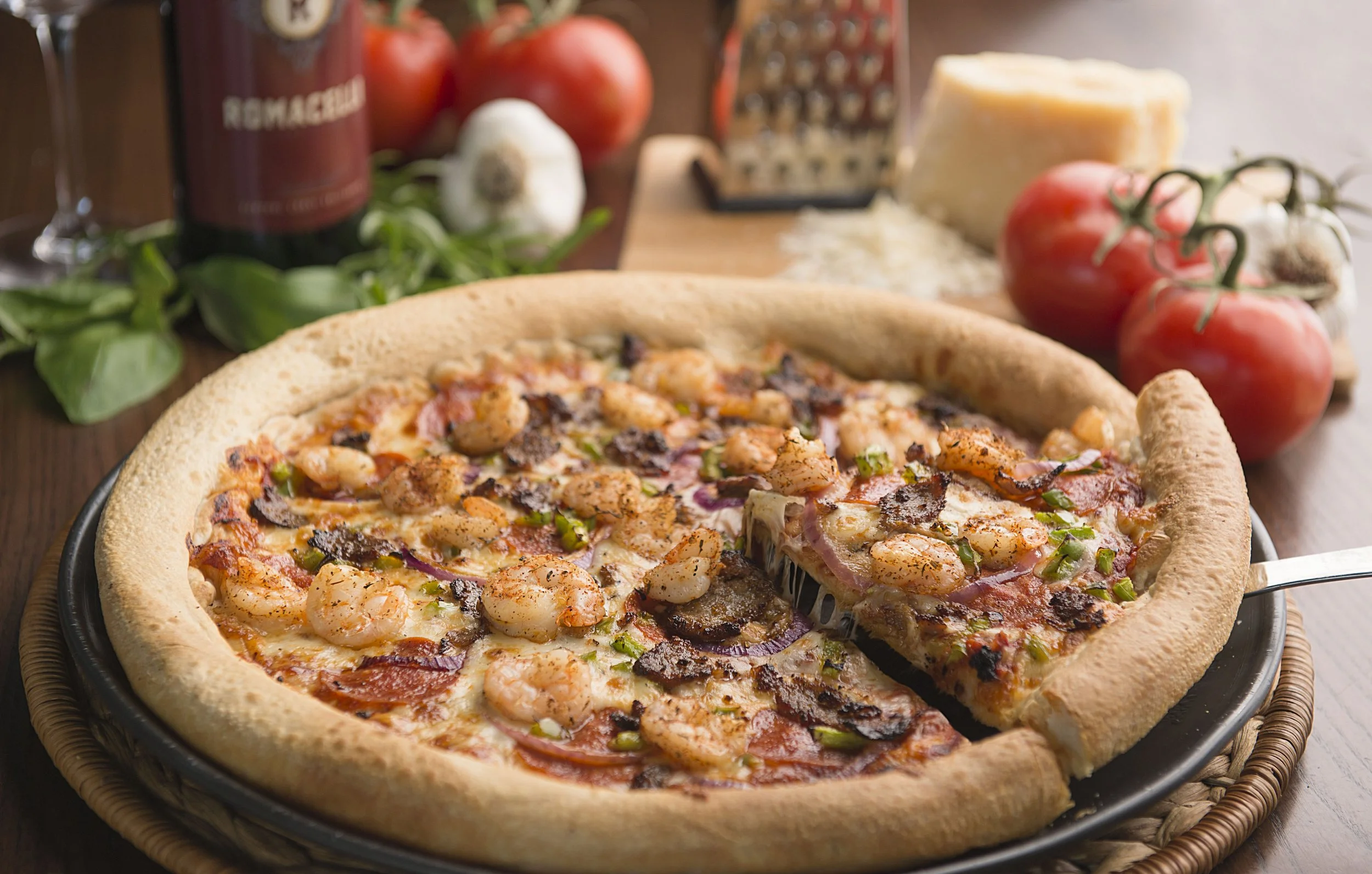 Close-up of a pizza topped with shrimp, sausage, red onions, and green peppers, set on a black pan with fresh tomatoes, garlic, cheese, and a grater in the background.