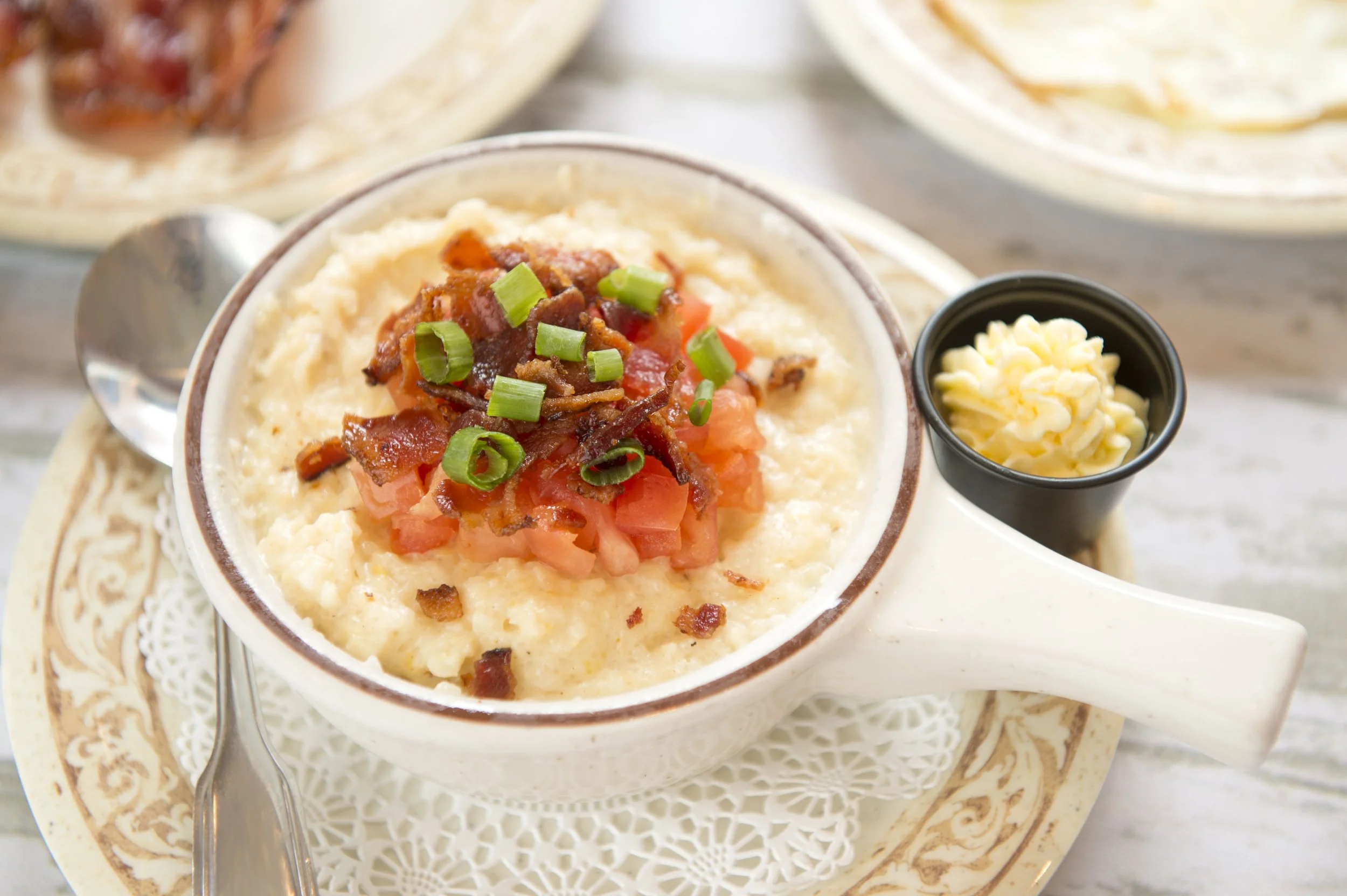 Bowl of grits topped with bacon, tomatoes, and green onions, served with a butter pat on the side.
