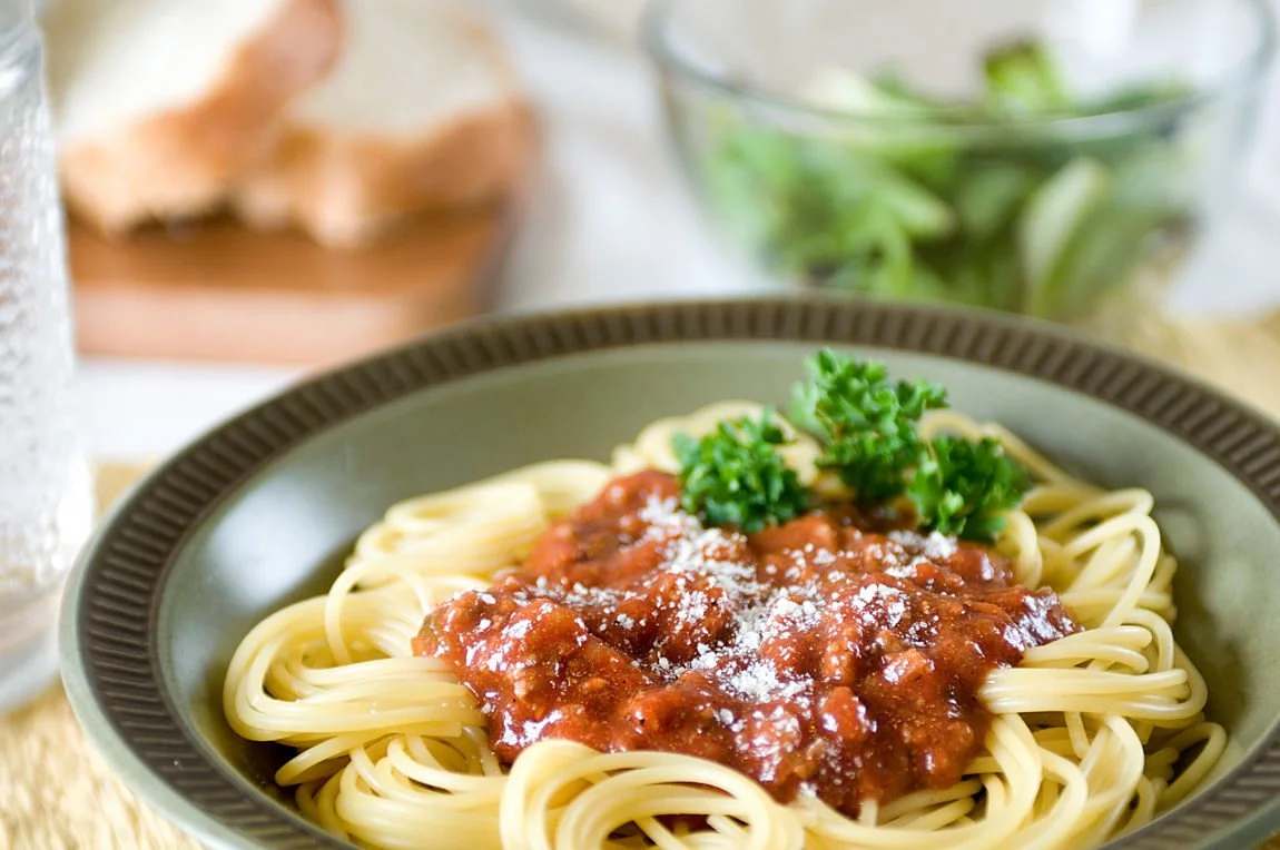 Plate of spaghetti with meat sauce and parsley garnish, table setting with bread and salad in the background.