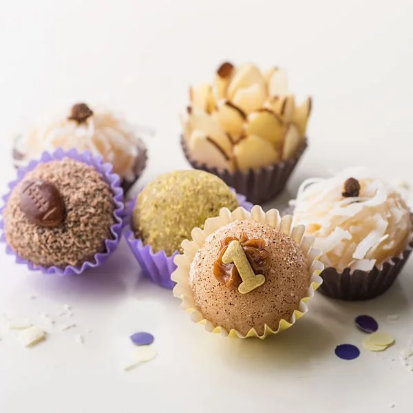 Assorted brigadeiros in colorful wrappers, topped with nuts, coconut, shaved chocolate, and decorative number one. Confectionery arranged with purple and white confetti on a light background.