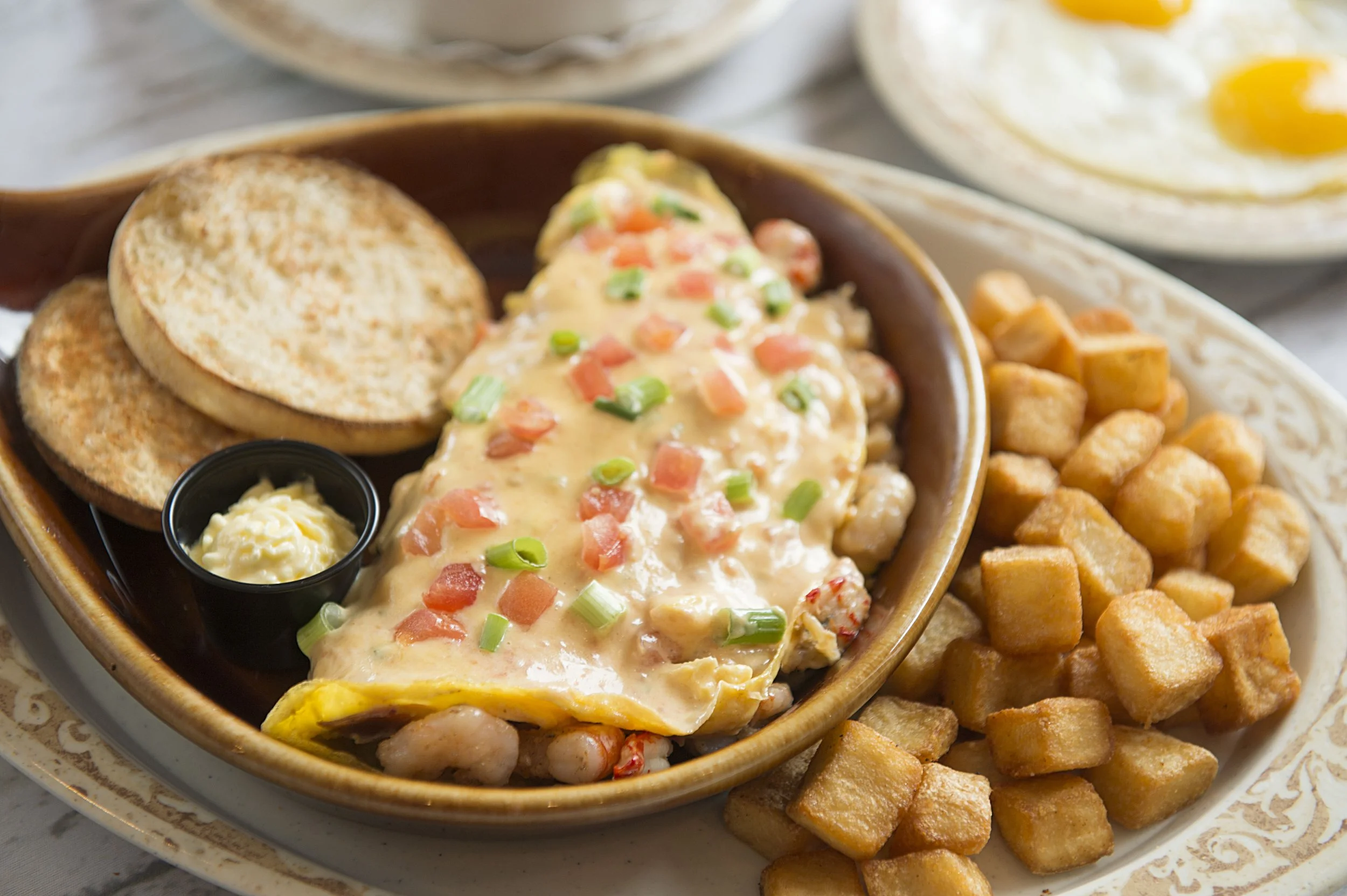 Breakfast platter with a loaded omelette topped with cheese, diced tomatoes, and green onions, served with toasted English muffins, butter on the side, and a portion of home fries; sunny-side-up eggs in the background.