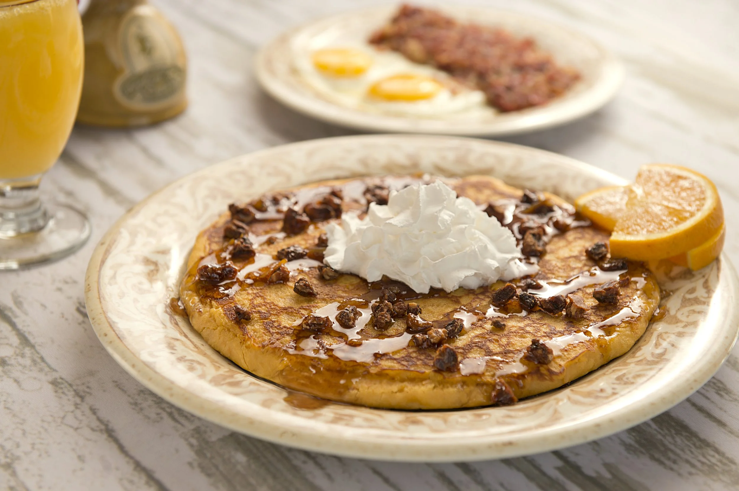 Pancake with whipped cream, chocolate chips, syrup, and orange slices; background shows eggs, bacon, and orange juice.