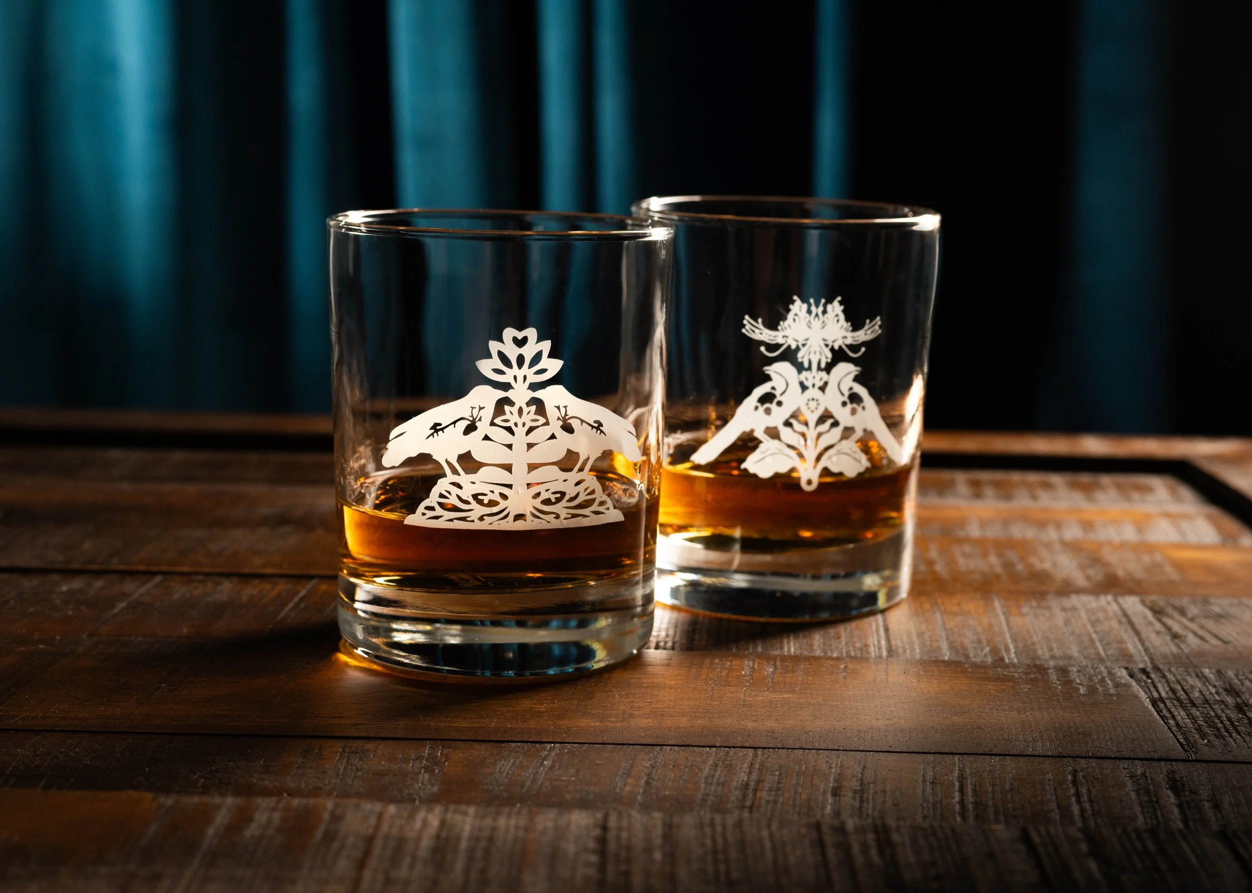 Two glasses of amber whiskey on a wooden table with decorative white designs, against a dark background.