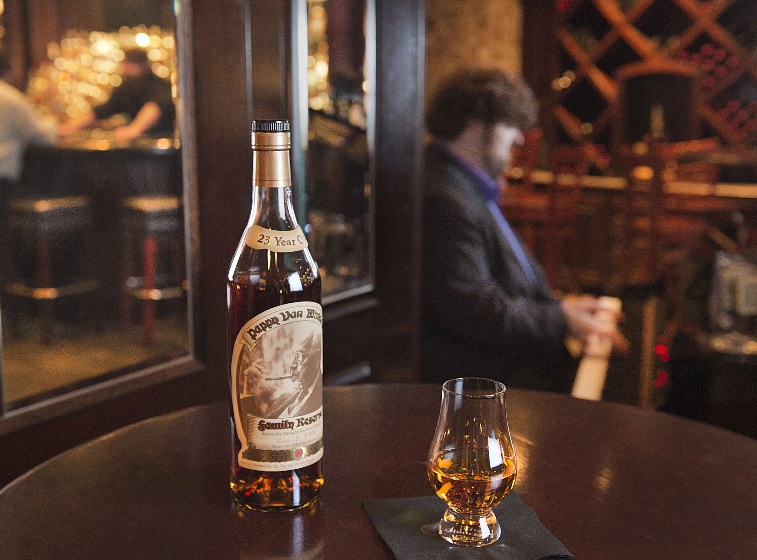 A bottle of 23-year-old Pappy Van Winkle bourbon on a table with a glass of bourbon, in a bar setting with blurred background showing a person playing piano.