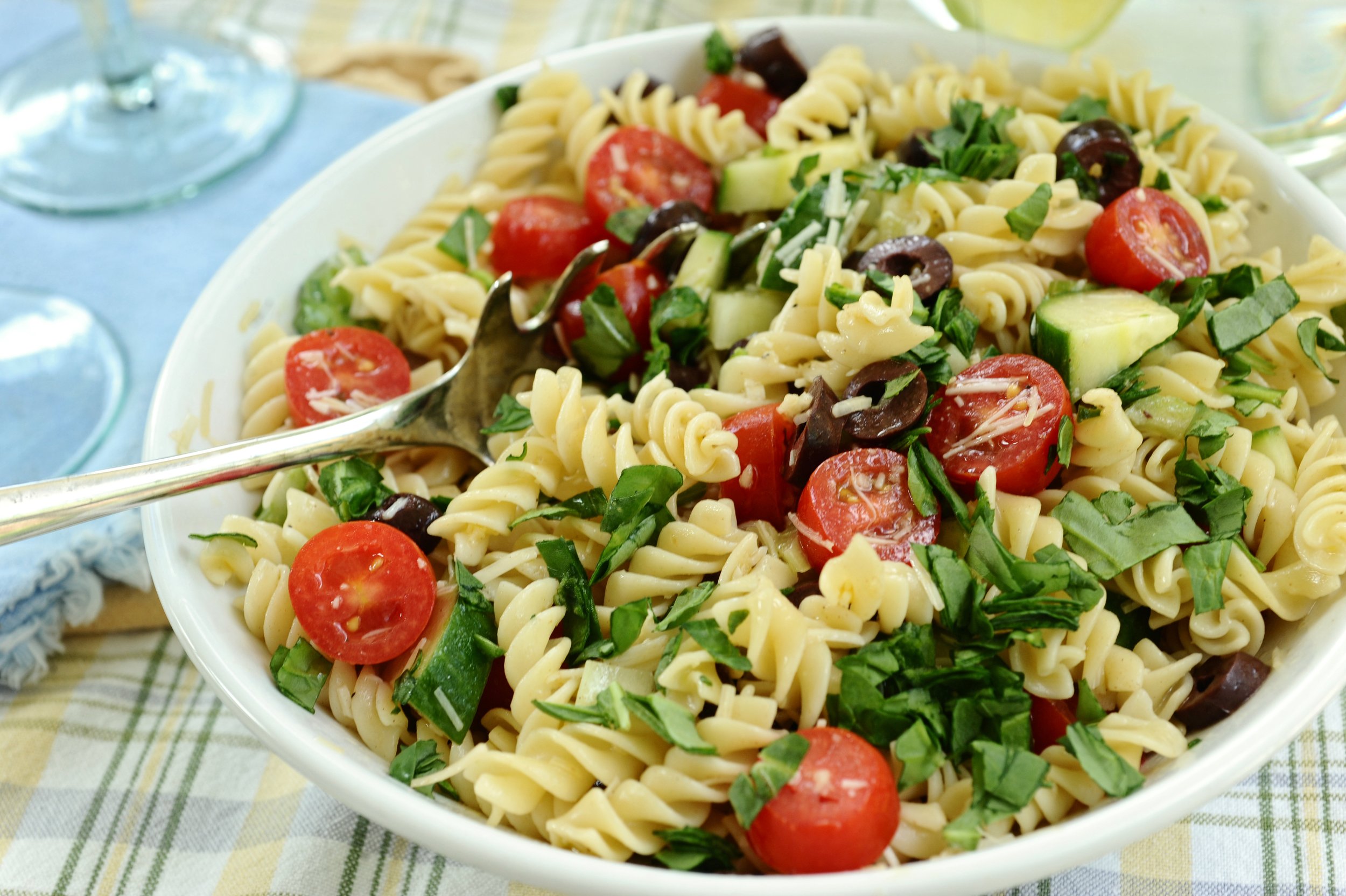 Bowl of pasta salad with rotini, cherry tomatoes, spinach, cucumbers, and black olives, served with a fork on a checkered tablecloth.
