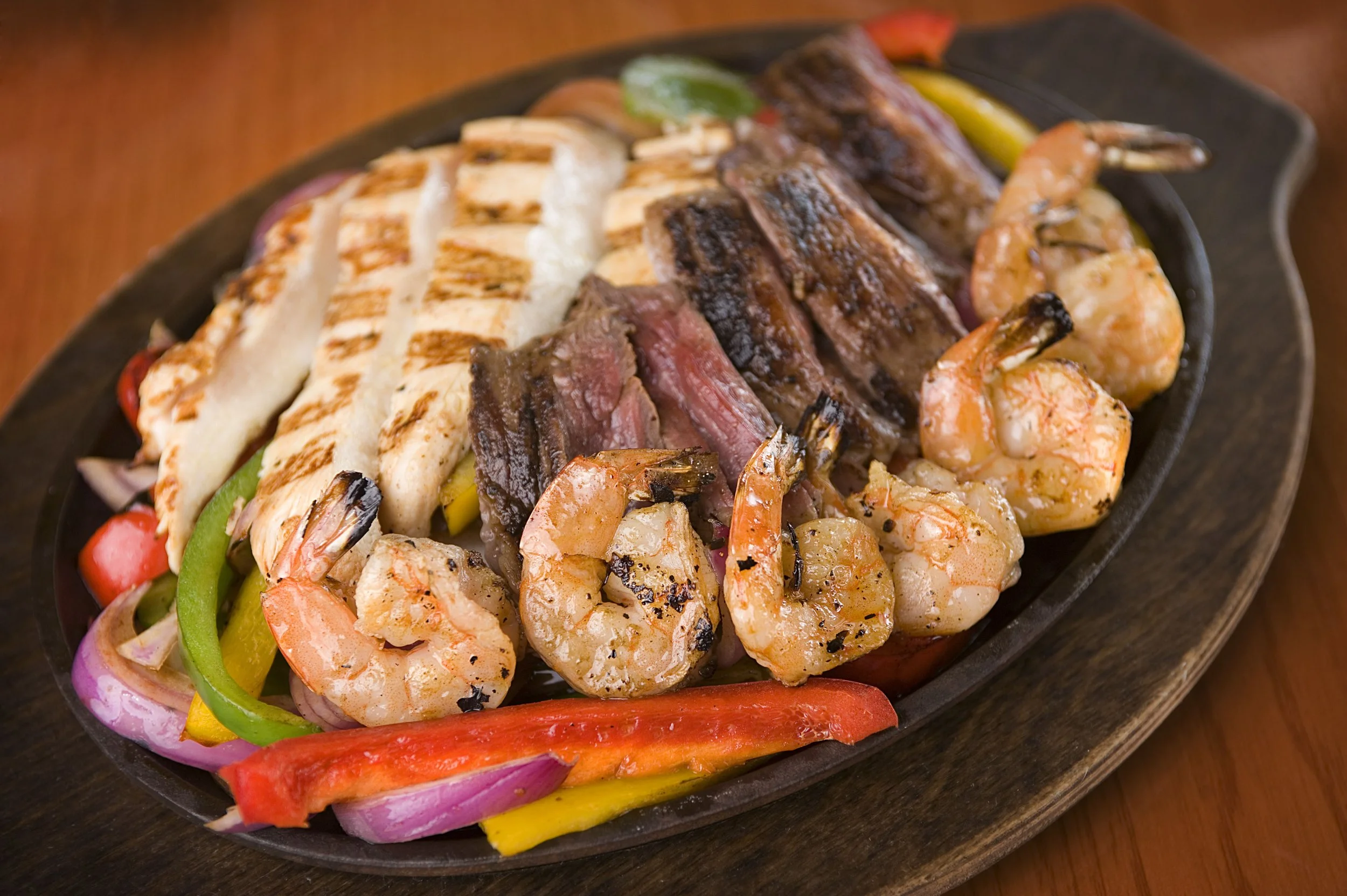 A sizzling platter of fajitas with grilled chicken, steak, shrimp, bell peppers, and onions, served on a wooden table.
