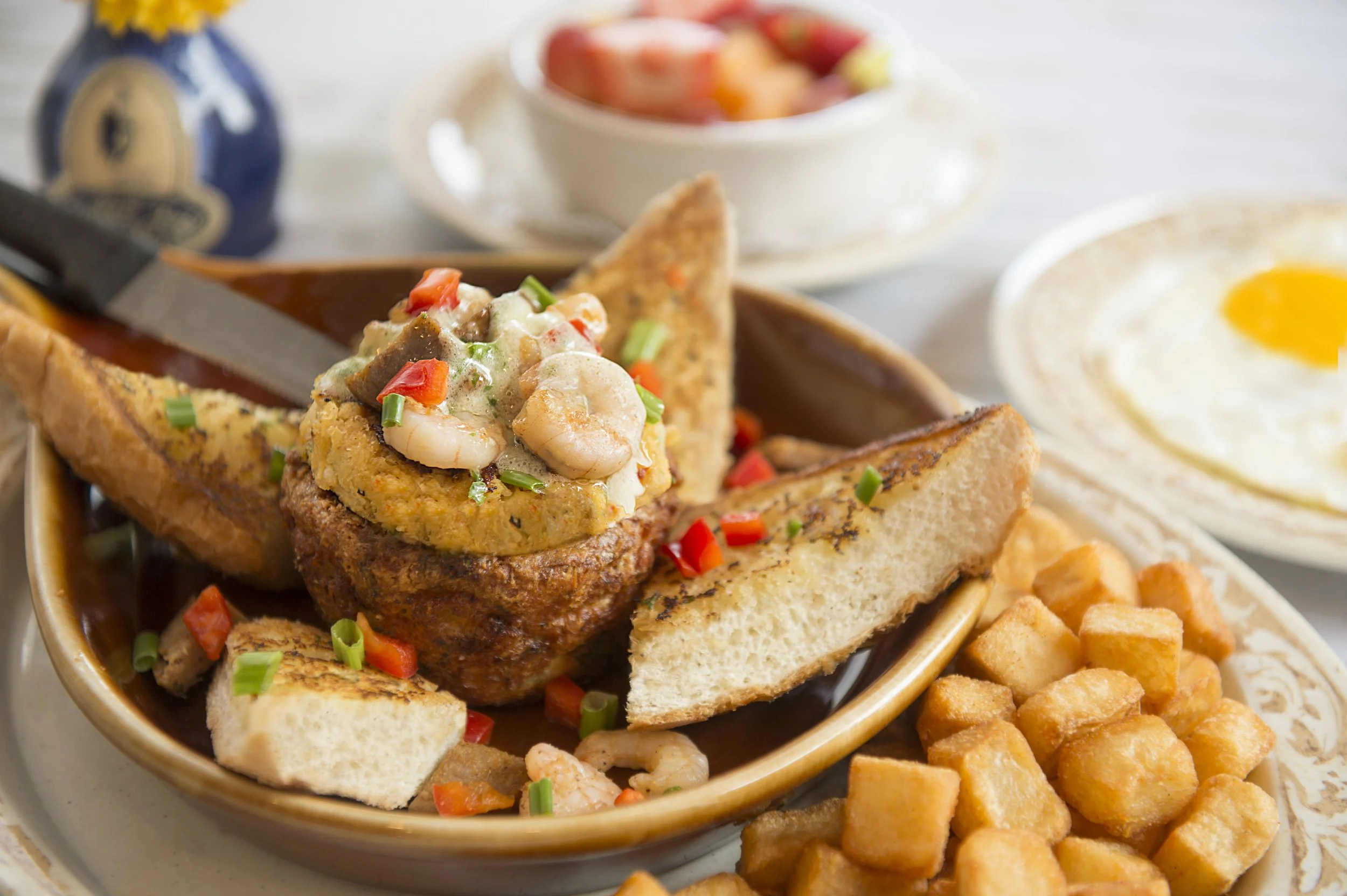 A close-up of a breakfast dish featuring French toast wedges with a savory topping, including shrimp, diced vegetables, and sauce, accompanied by crispy diced potatoes. In the background, there's a bowl of fruit salad and a plate with a sunny-side-up