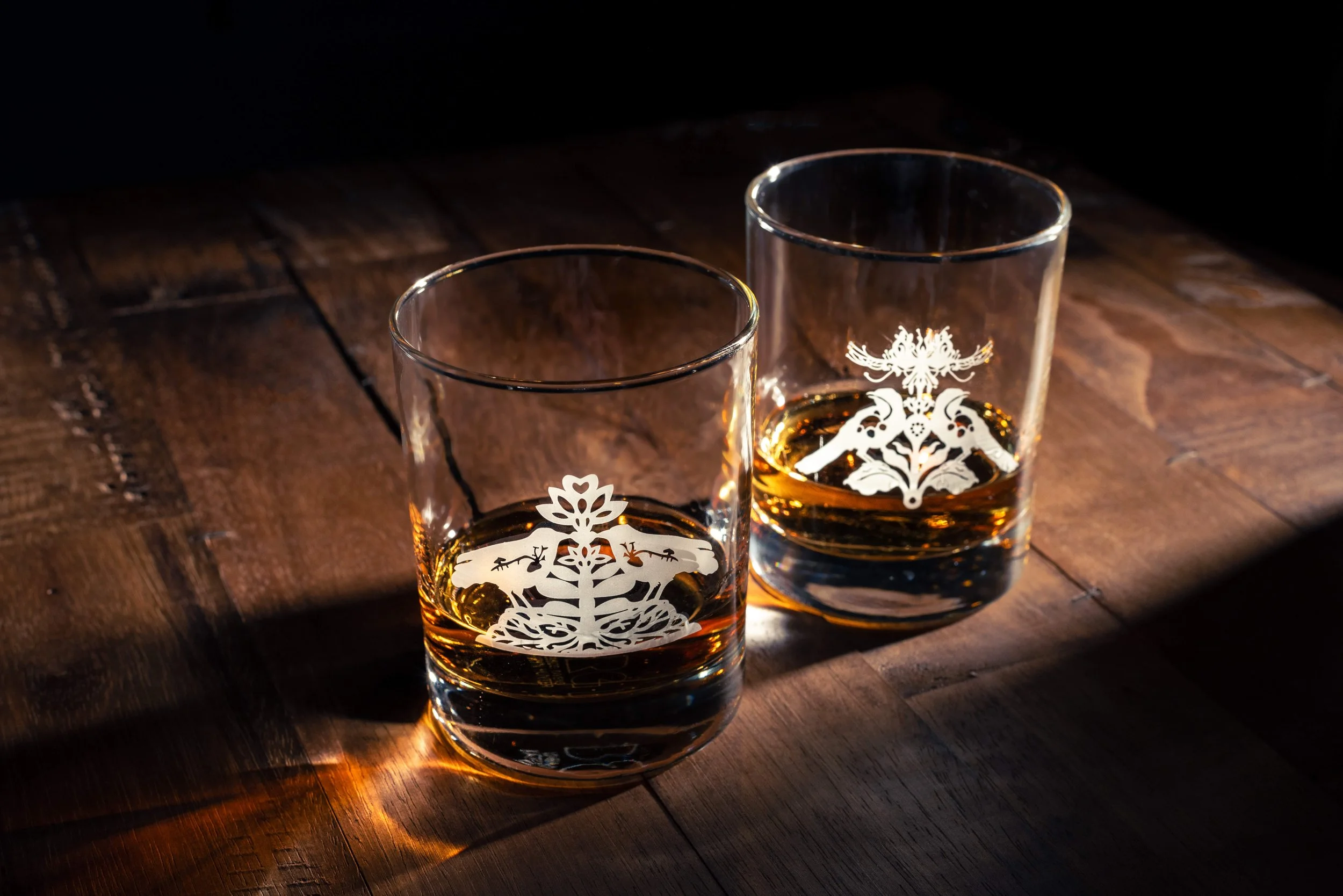 Two whiskey glasses with intricate white designs, partially filled with whiskey, on a wooden table.