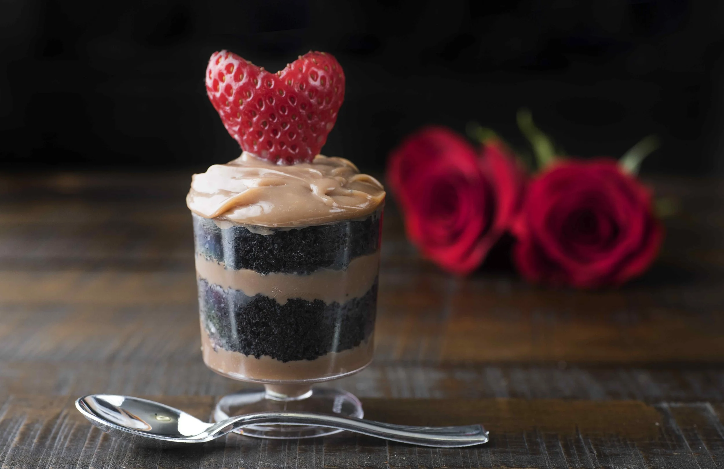 Dessert parfait with layers of chocolate cake and cream, topped with a heart-shaped strawberry, beside two red roses on a wooden table.